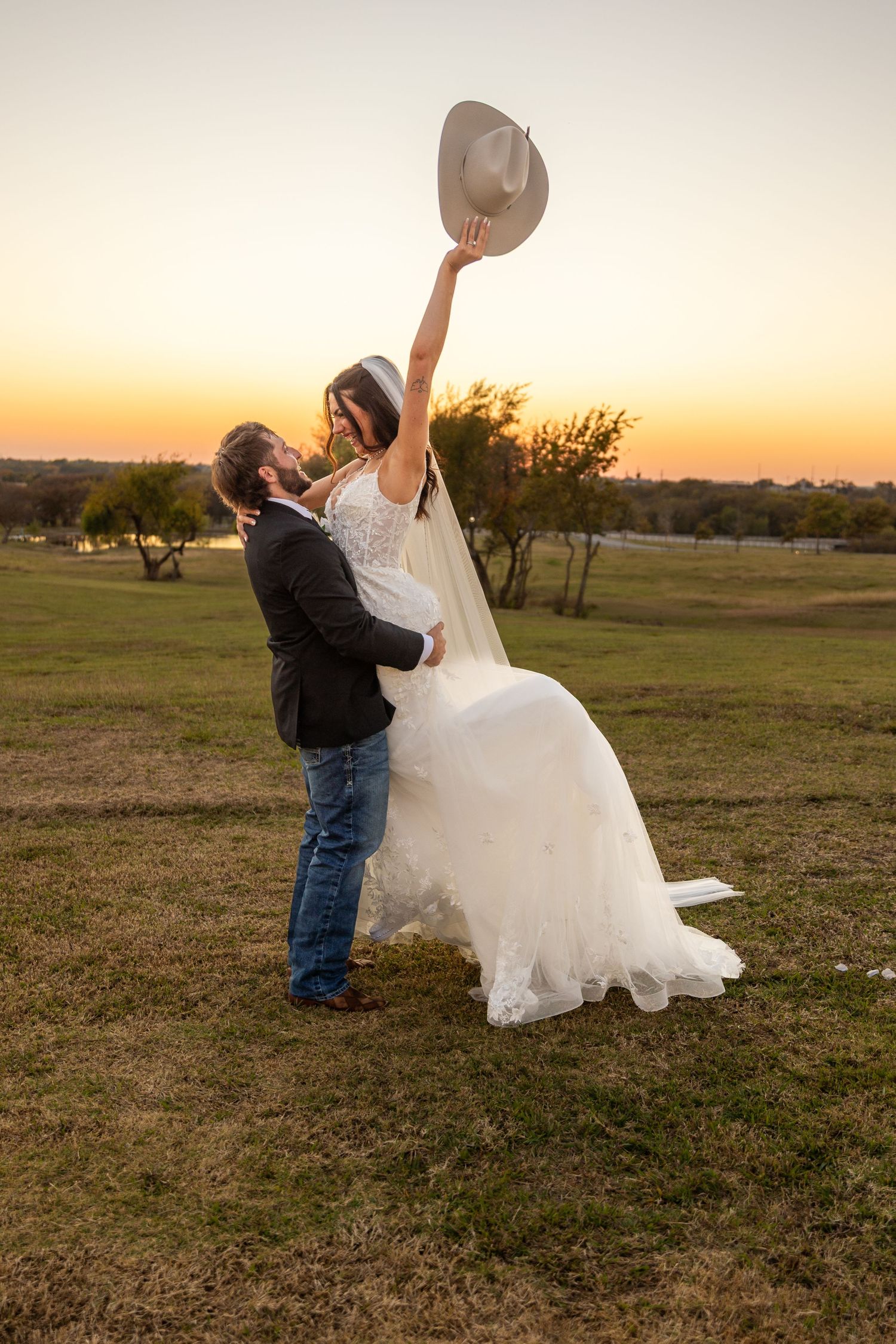 Playful wedding photo shoot in a field at golden hour with flowing white dress and cowboy hat.