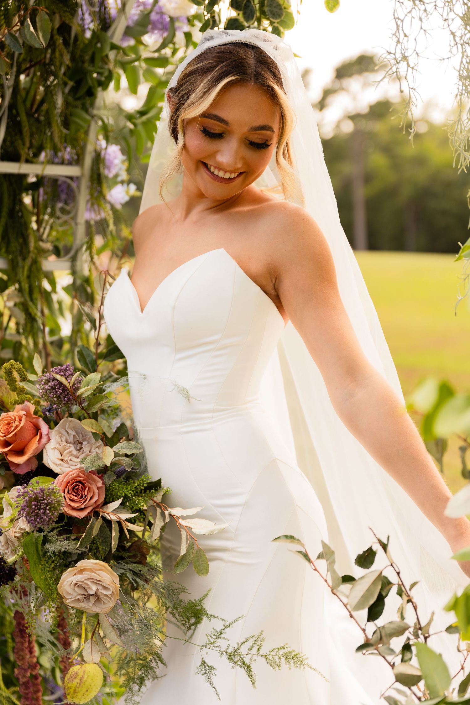 Bride in strapless wedding gown poses with floral arrangement against natural backdrop.