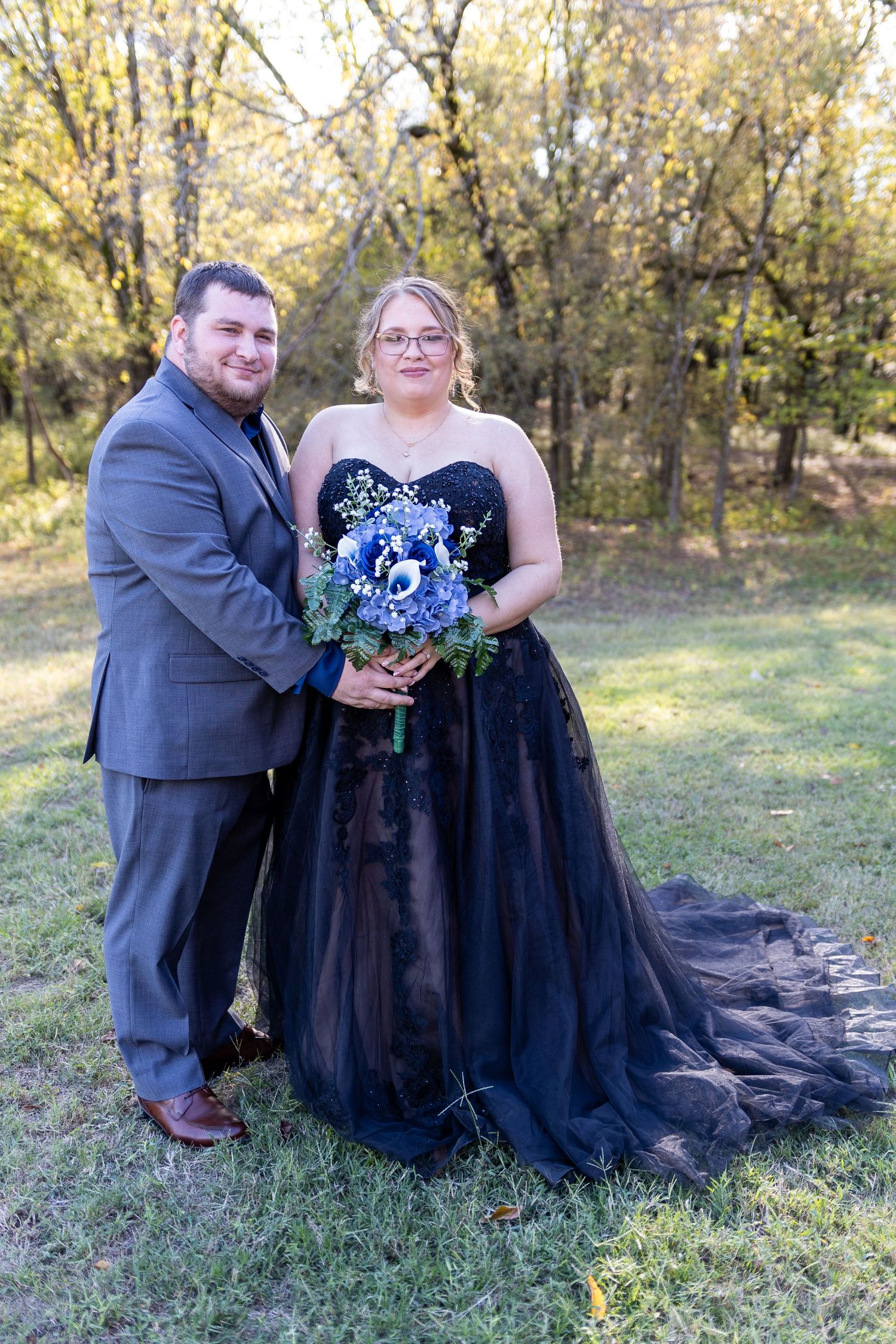 Couple in formal wear sitting on grass with blue bouquet during autumn photo session.