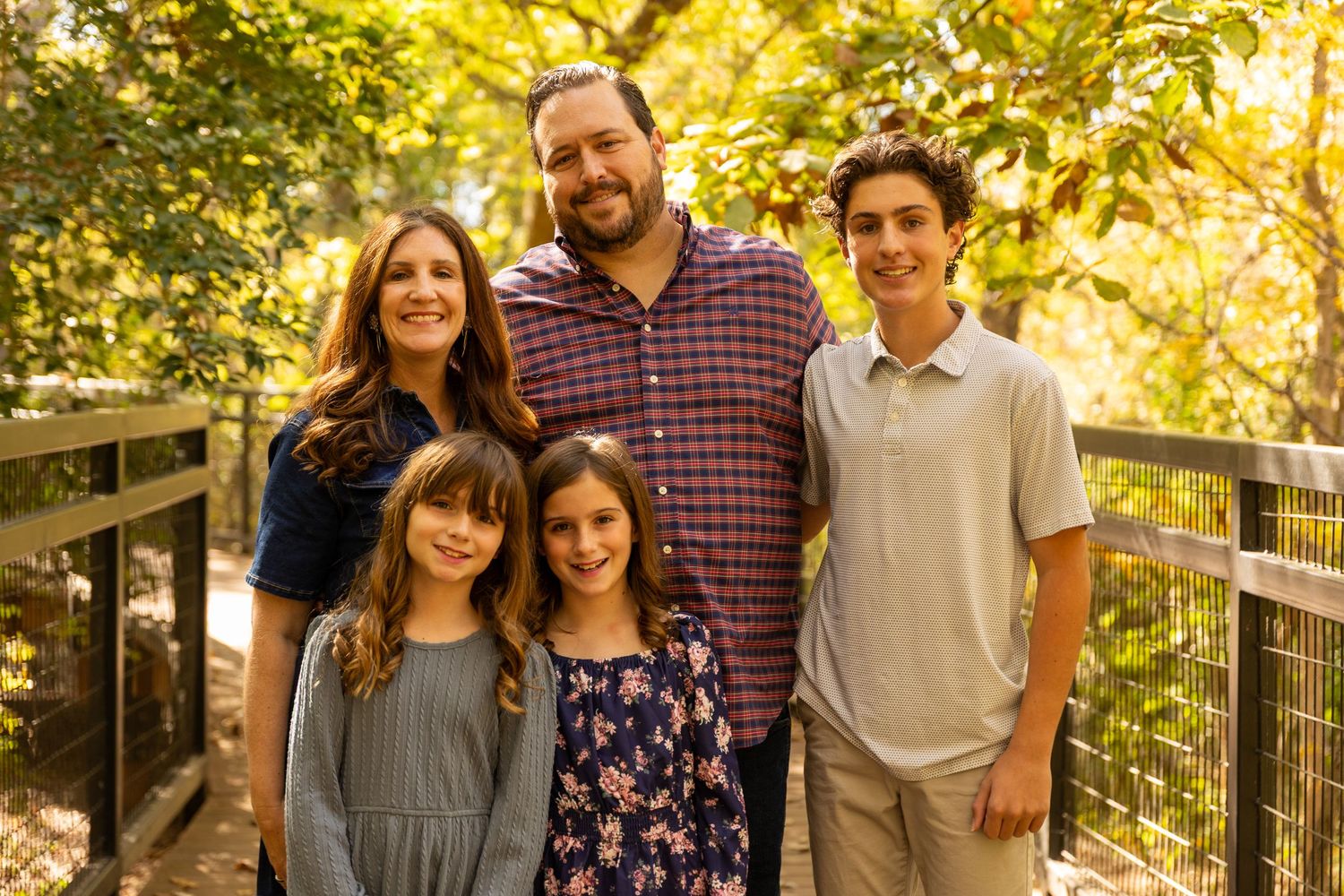 Family portrait on wooden bridge surrounded by golden autumn foliage in outdoor nature setting.