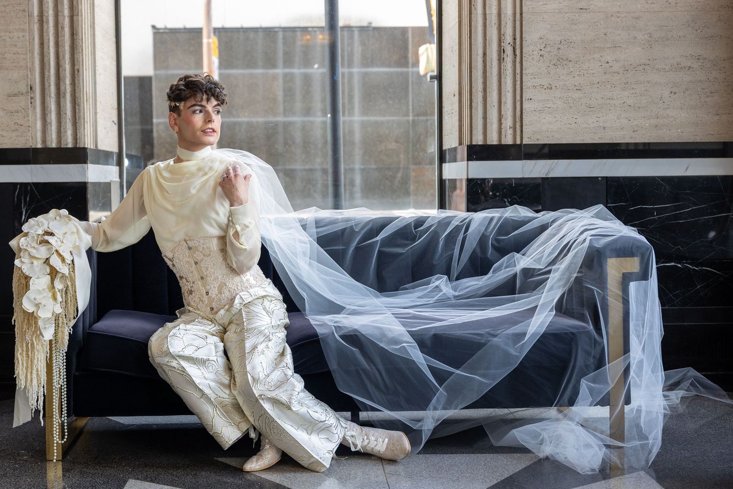 Person in white avant-garde outfit reclines on dark sofa with flowing tulle fabric in elegant interior setting.