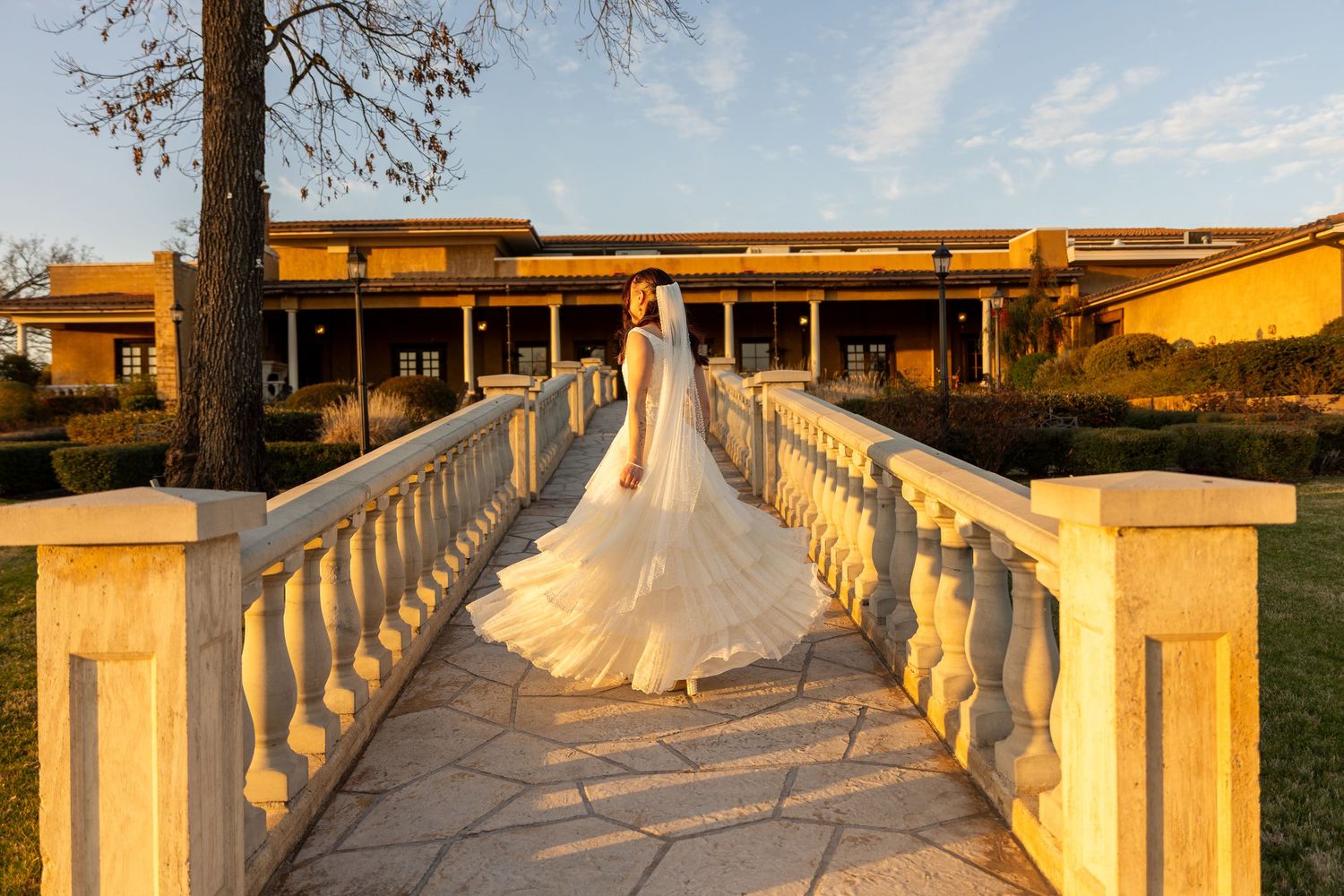 Bride in flowing white gown stands on elegant stone bridge at golden hour outside a yellow venue.