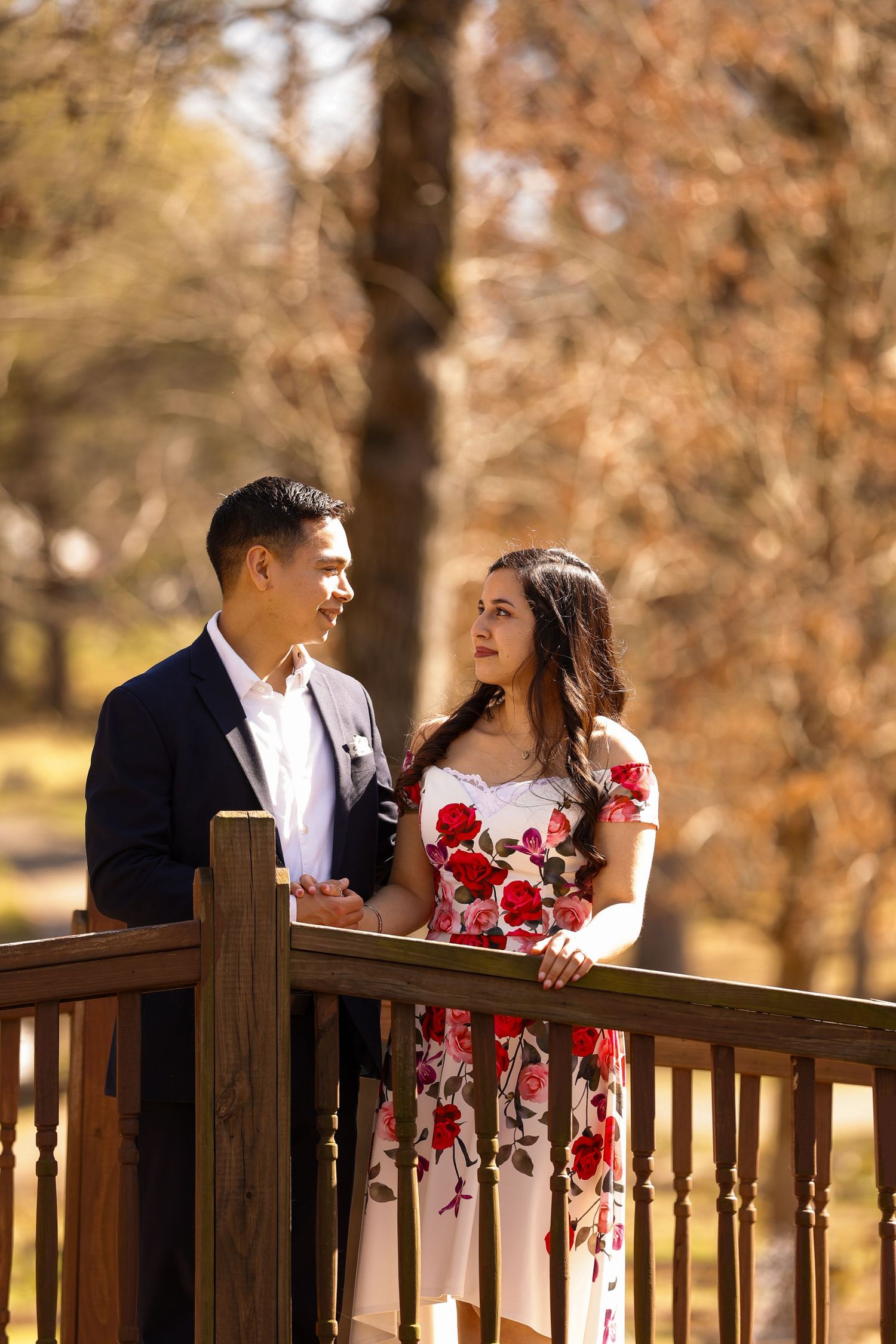 A couple shares a loving glance while standing on a wooden deck in autumn sunlight.