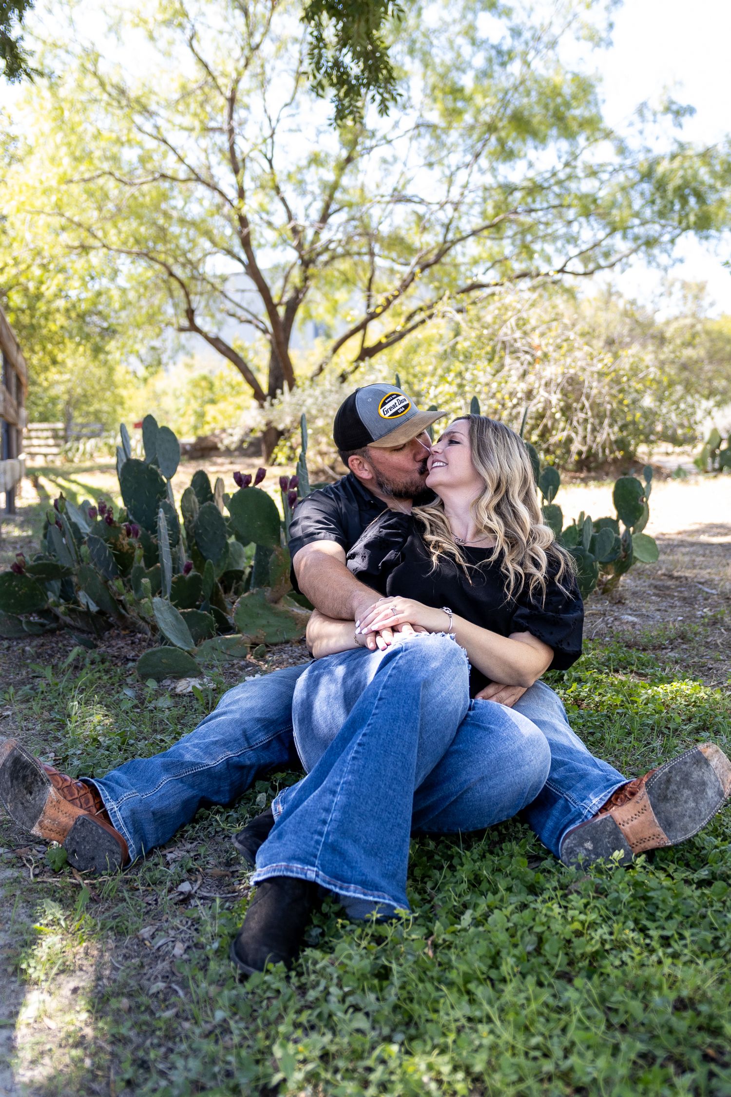 A couple shares intimate moments while lying in grass near cacti during an outdoor photoshoot.