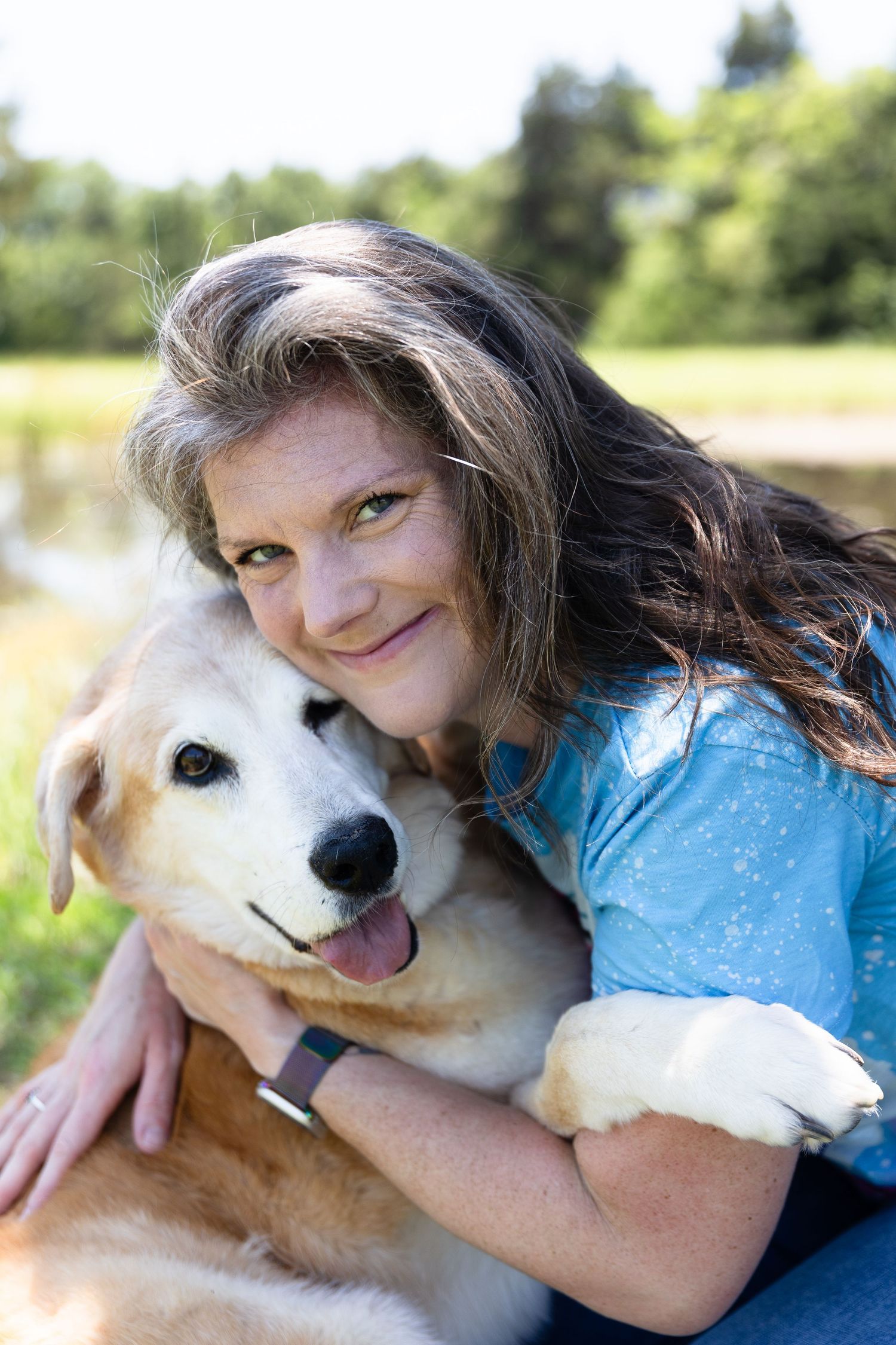 A happy golden retriever dog being hugged and cuddled outdoors next to a lake on a sunny day.