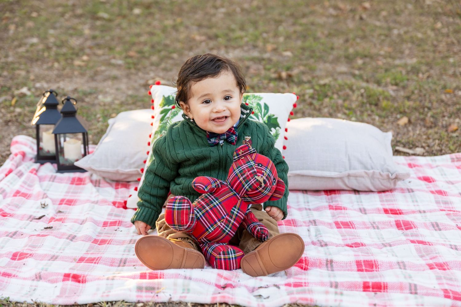 A toddler in winter clothing sits on a red checkered picnic blanket with decorative pillows outdoors.