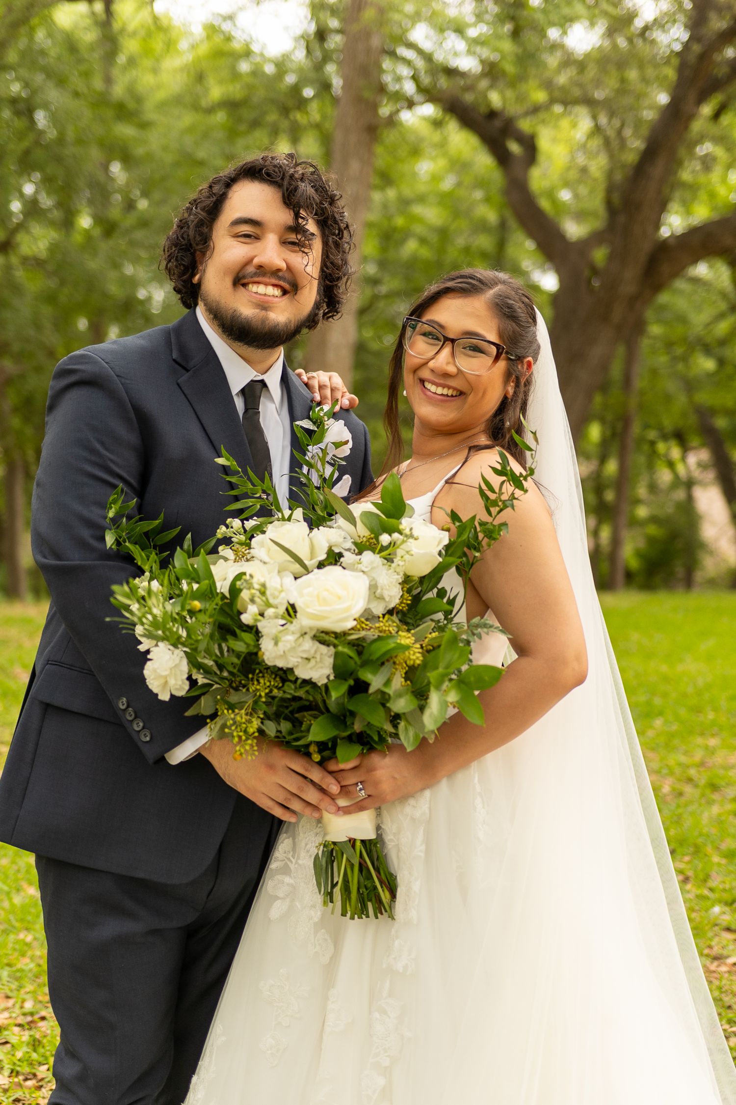 A joyful wedding couple poses with a beautiful white and green floral bouquet in an outdoor garden setting.