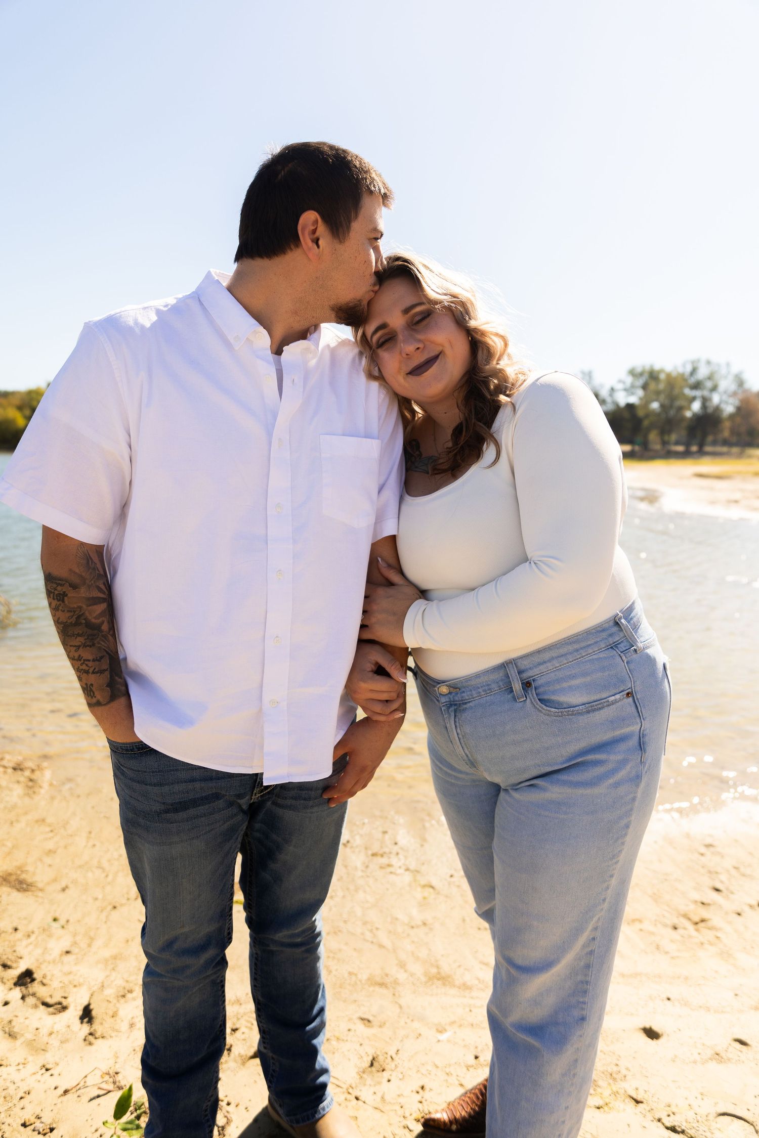 Couple embracing on a sunny beach wearing white and denim in casual romantic poses.