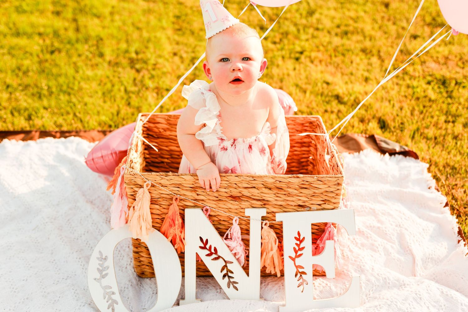 Baby in pink dress plays near wicker basket on white blanket during sunset.