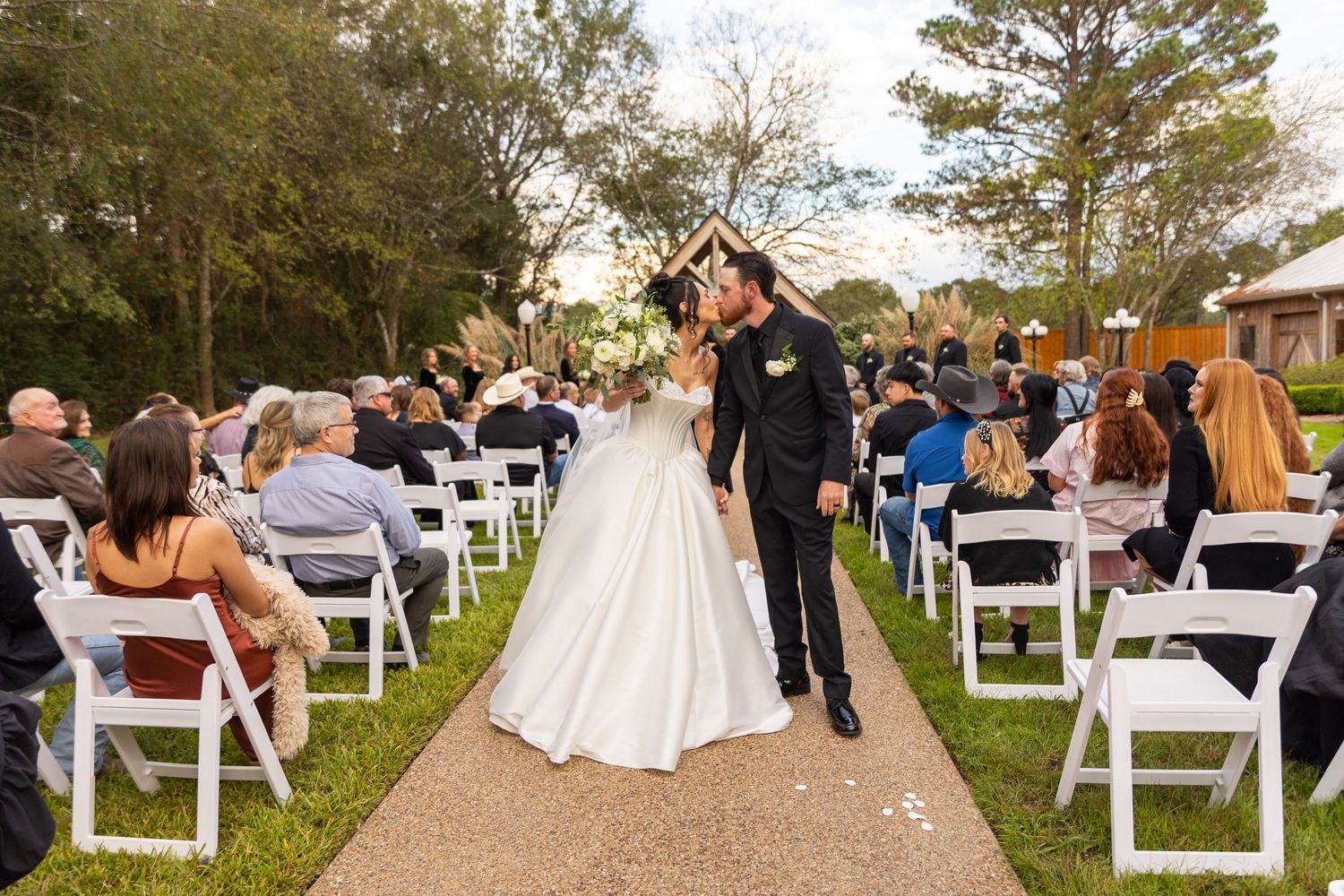 Outdoor wedding ceremony setup with white chairs and guests arriving on a garden path.