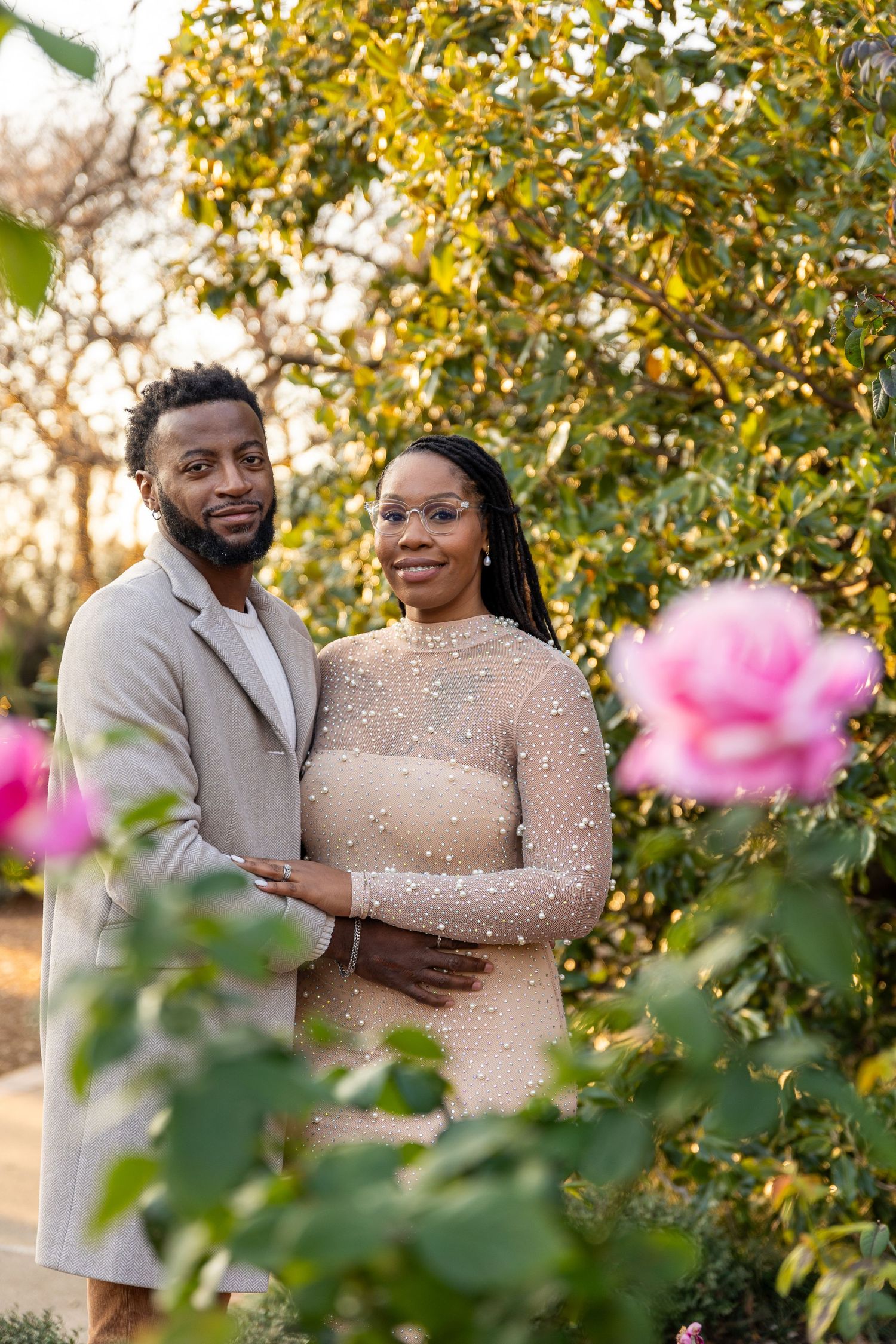 A couple in elegant beige outfits poses among blooming flowers in a garden setting.