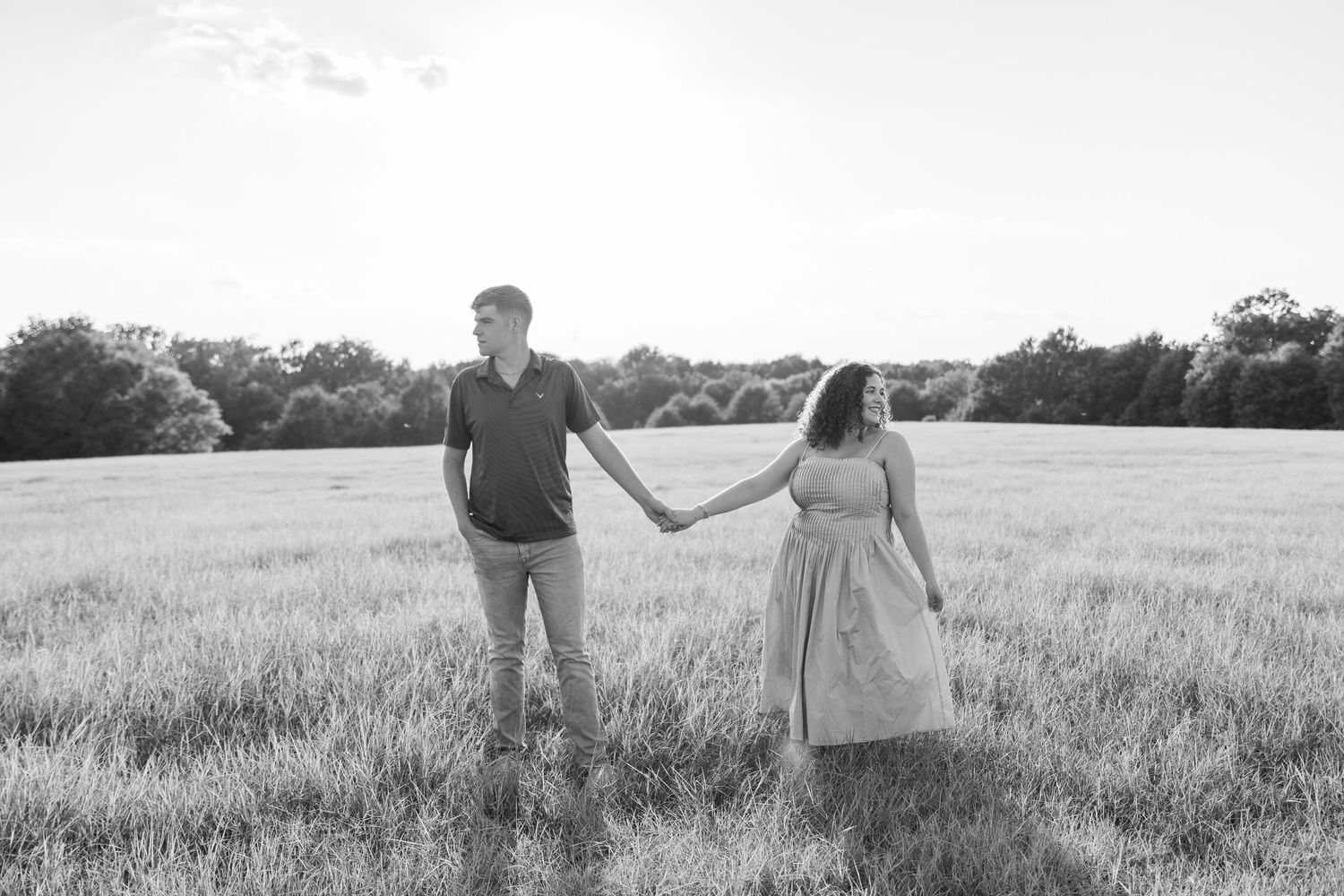 A romantic couple holds hands while standing in a sunlit meadow, captured in black and white.