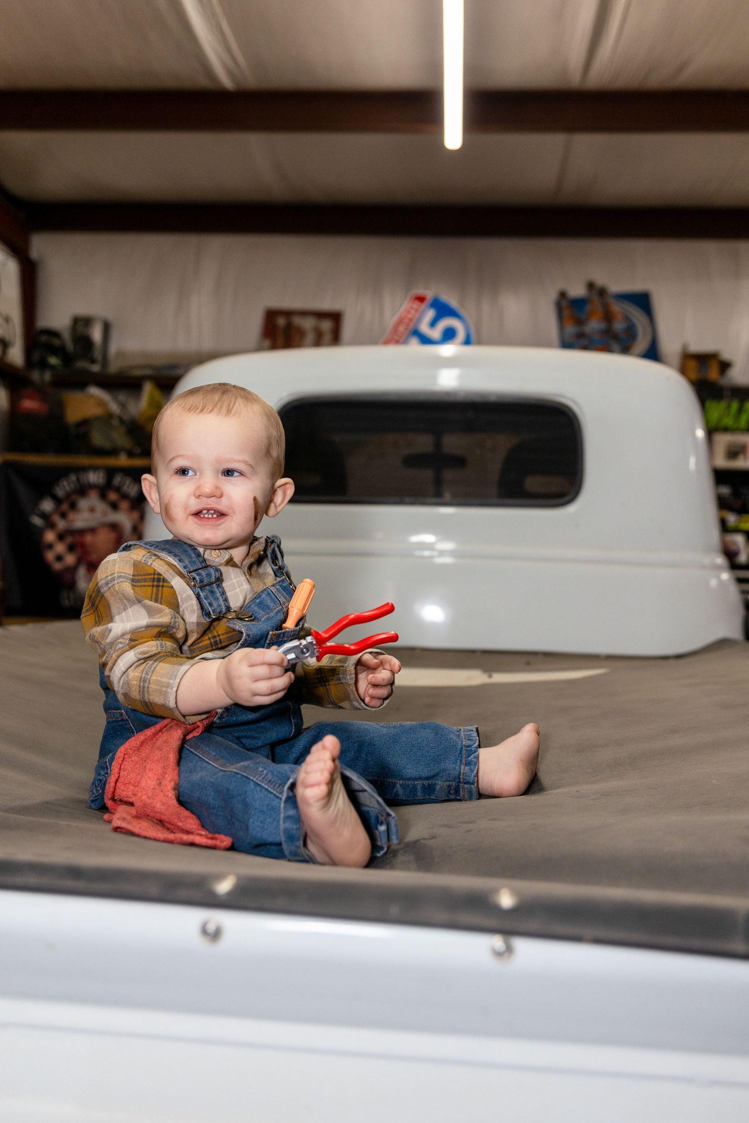 Baby in denim overalls plays with red tools on white vintage car hood.