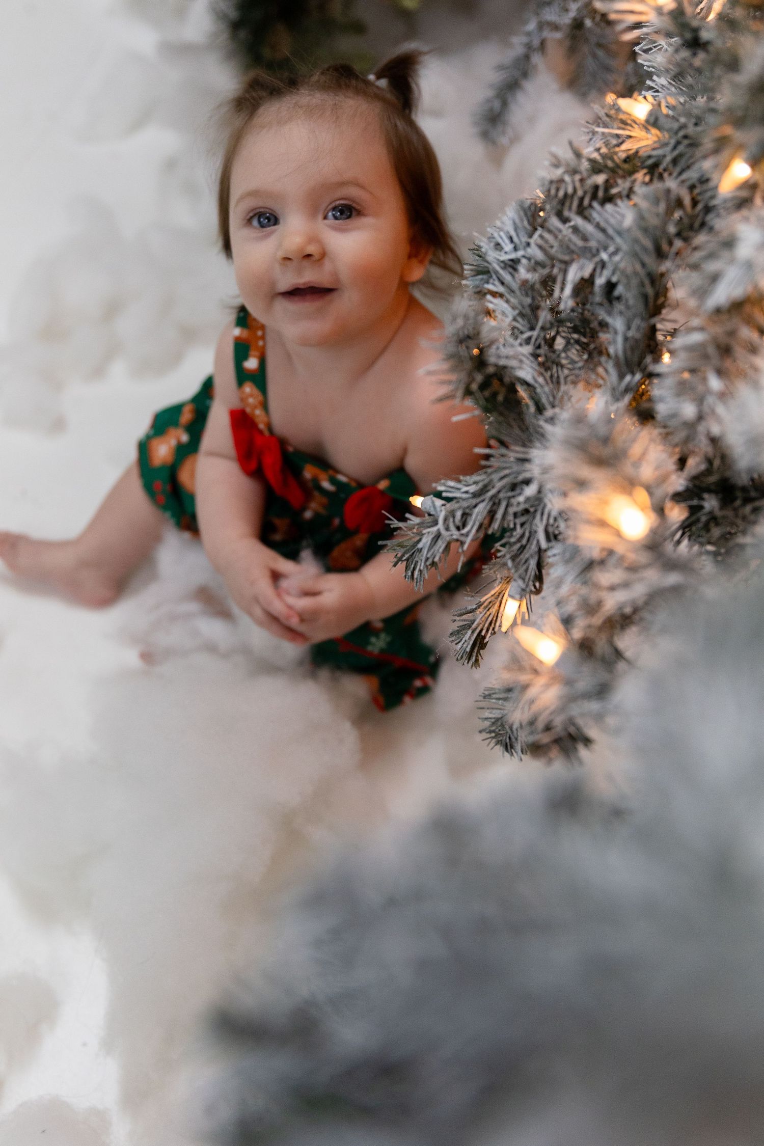 Baby in festive green and red outfit peeks through illuminated Christmas wreath decorations.
