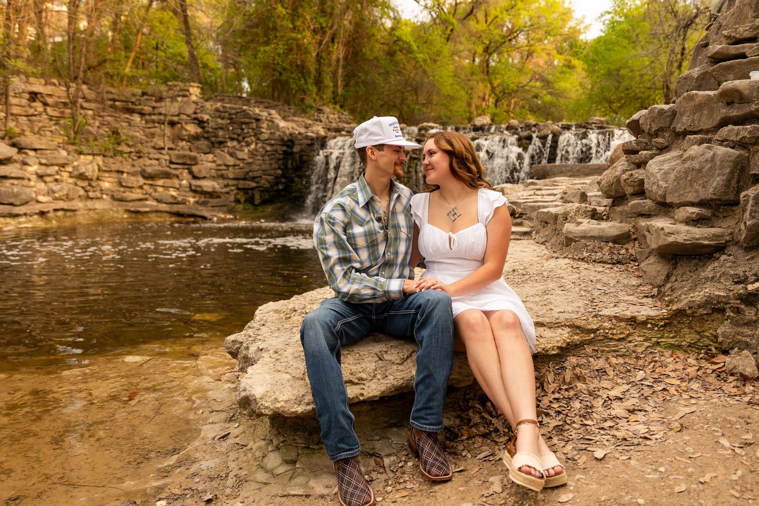 A couple sits together on rocks near a scenic waterfall in casual western attire.