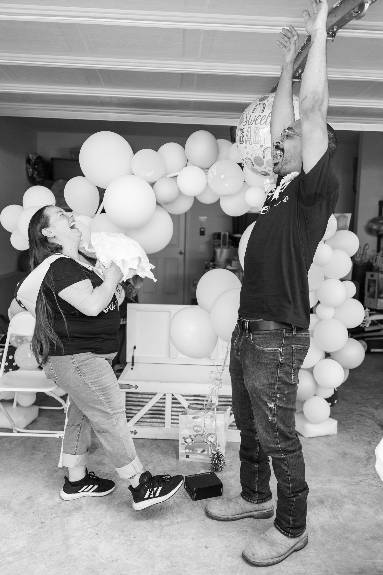 Black and white photo of people decorating with white balloon arch in garage.