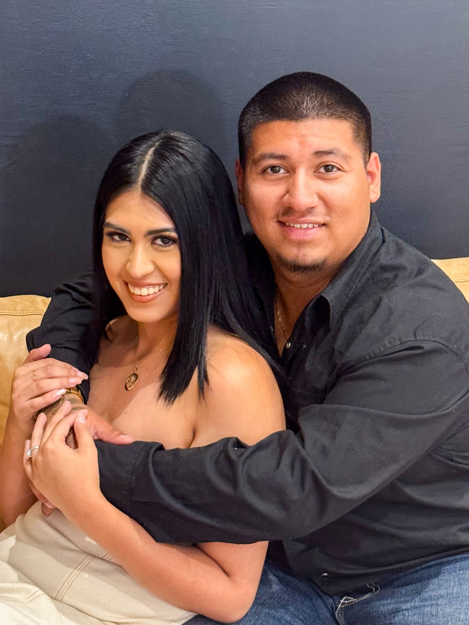 Couple in black attire posing together on beige couch for portrait.