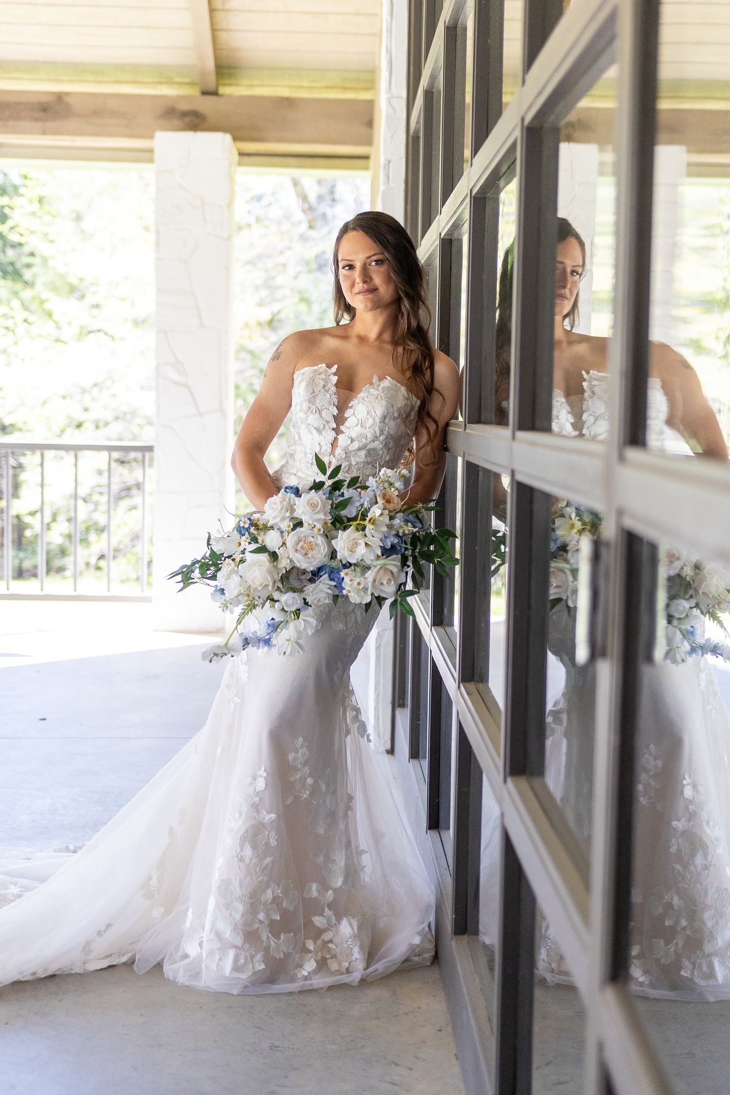 A bride in a lace wedding gown poses with her bouquet on a covered porch with window reflections.