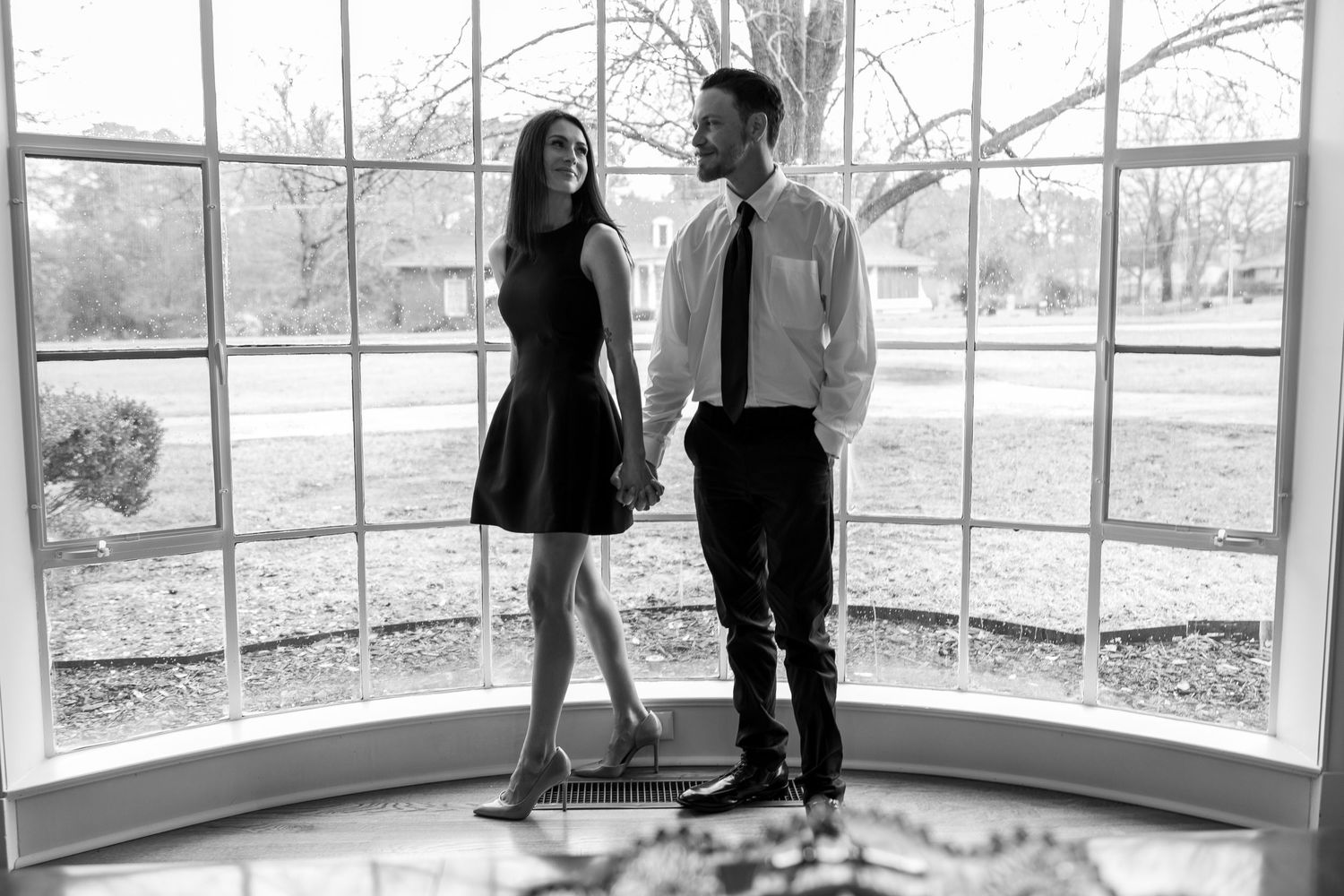 A black and white photo of a couple standing in front of large bay windows in formal attire.
