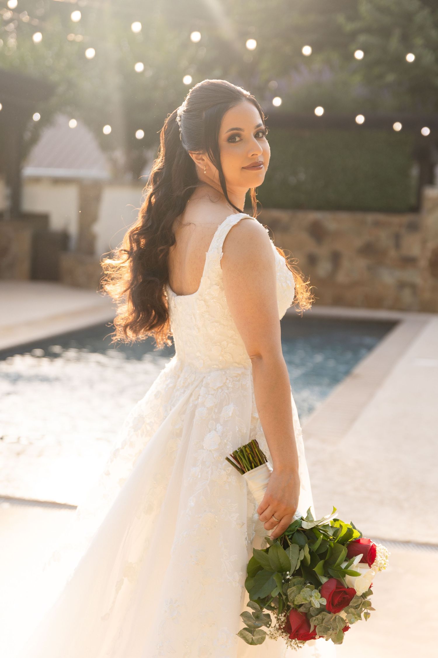 Bride in flowing white gown holds red and green bouquet by poolside with string lights overhead.