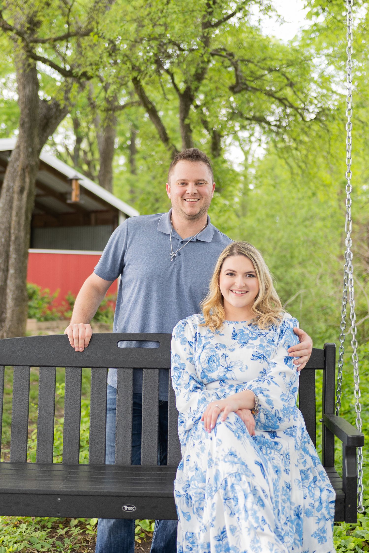 A couple sits together on a wooden park bench surrounded by green trees.