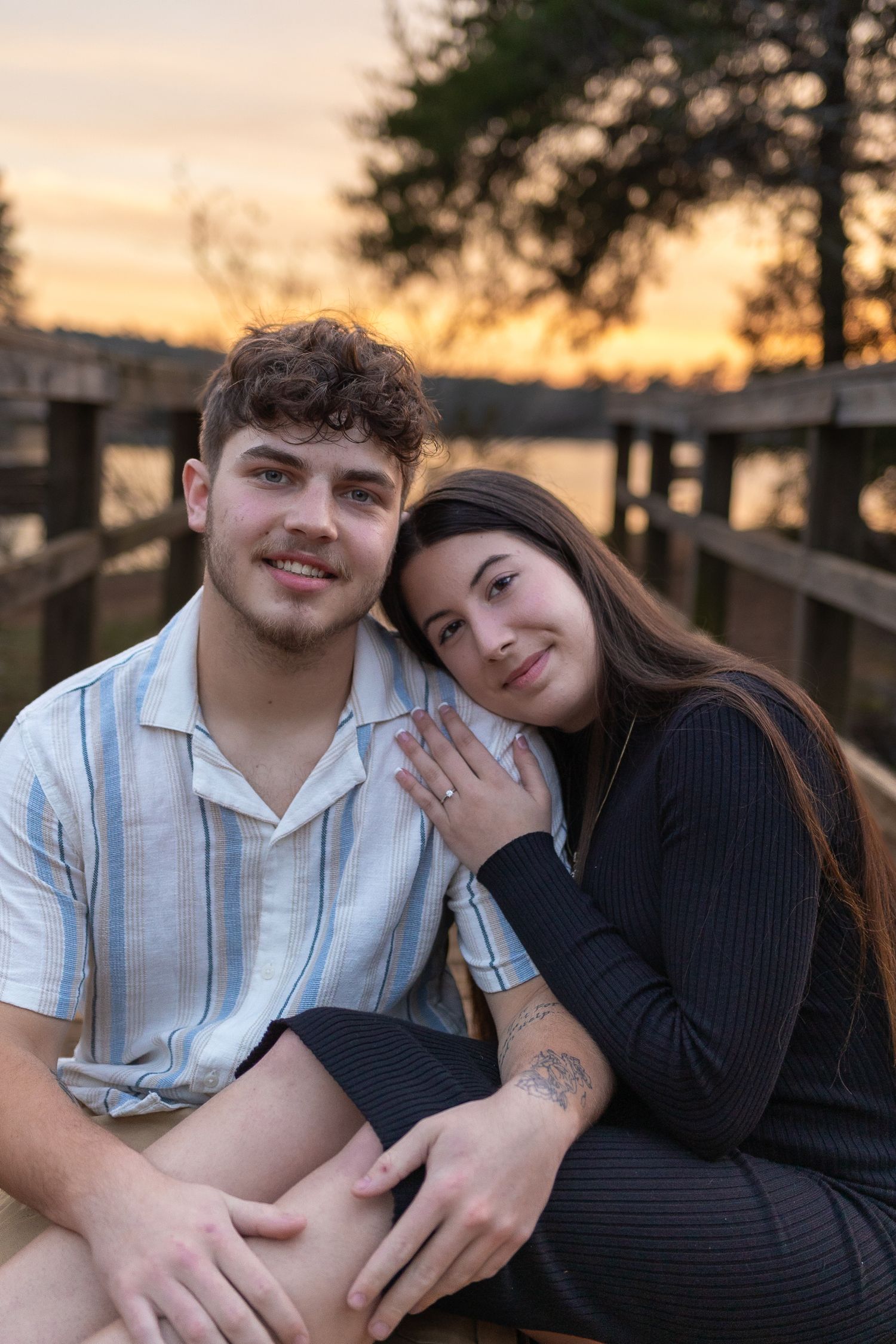A romantic couple poses together at sunset on a wooden deck, showing affection and closeness.