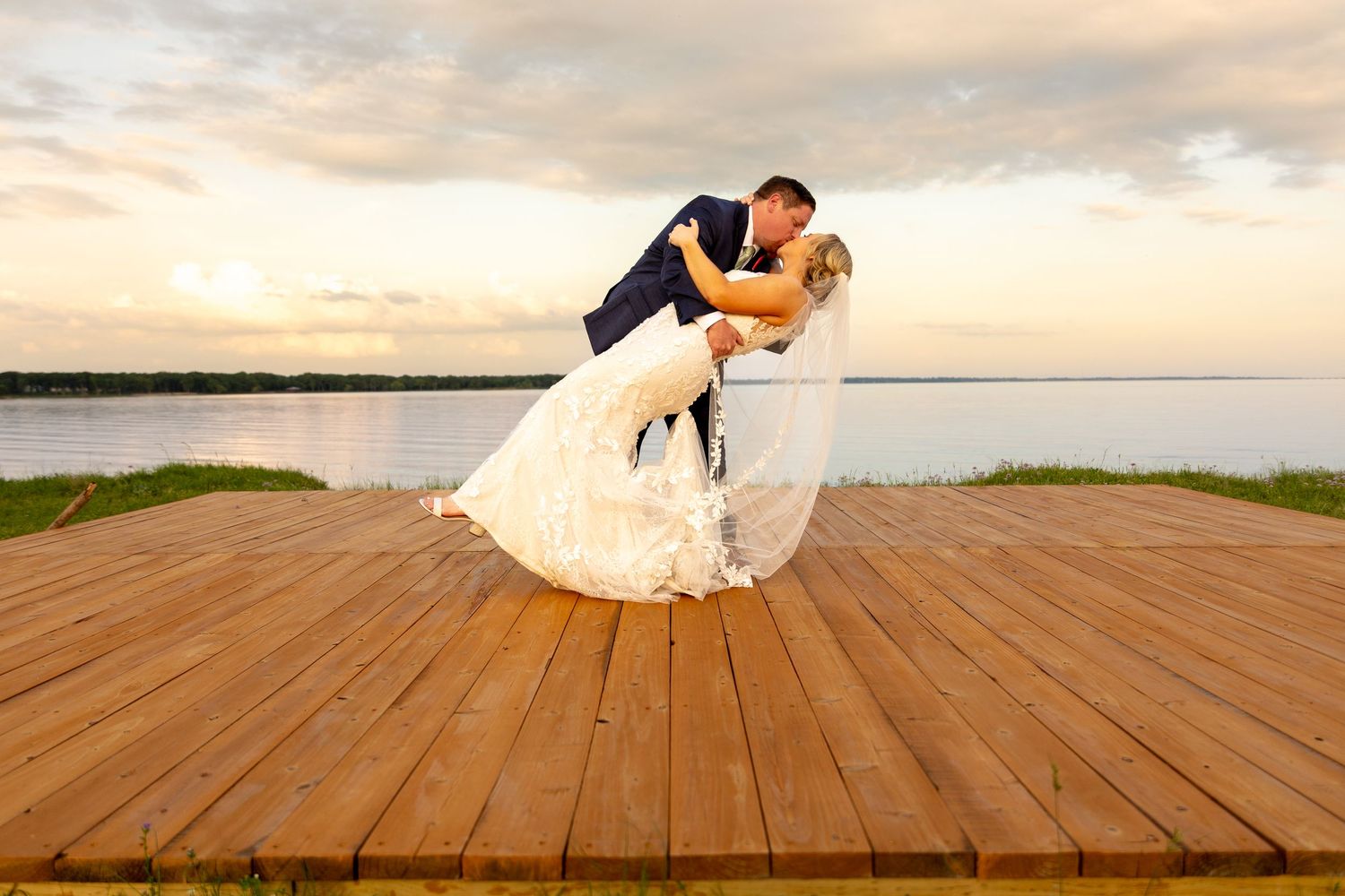 Wedding couple performs dramatic dip pose on wooden deck at sunset.
