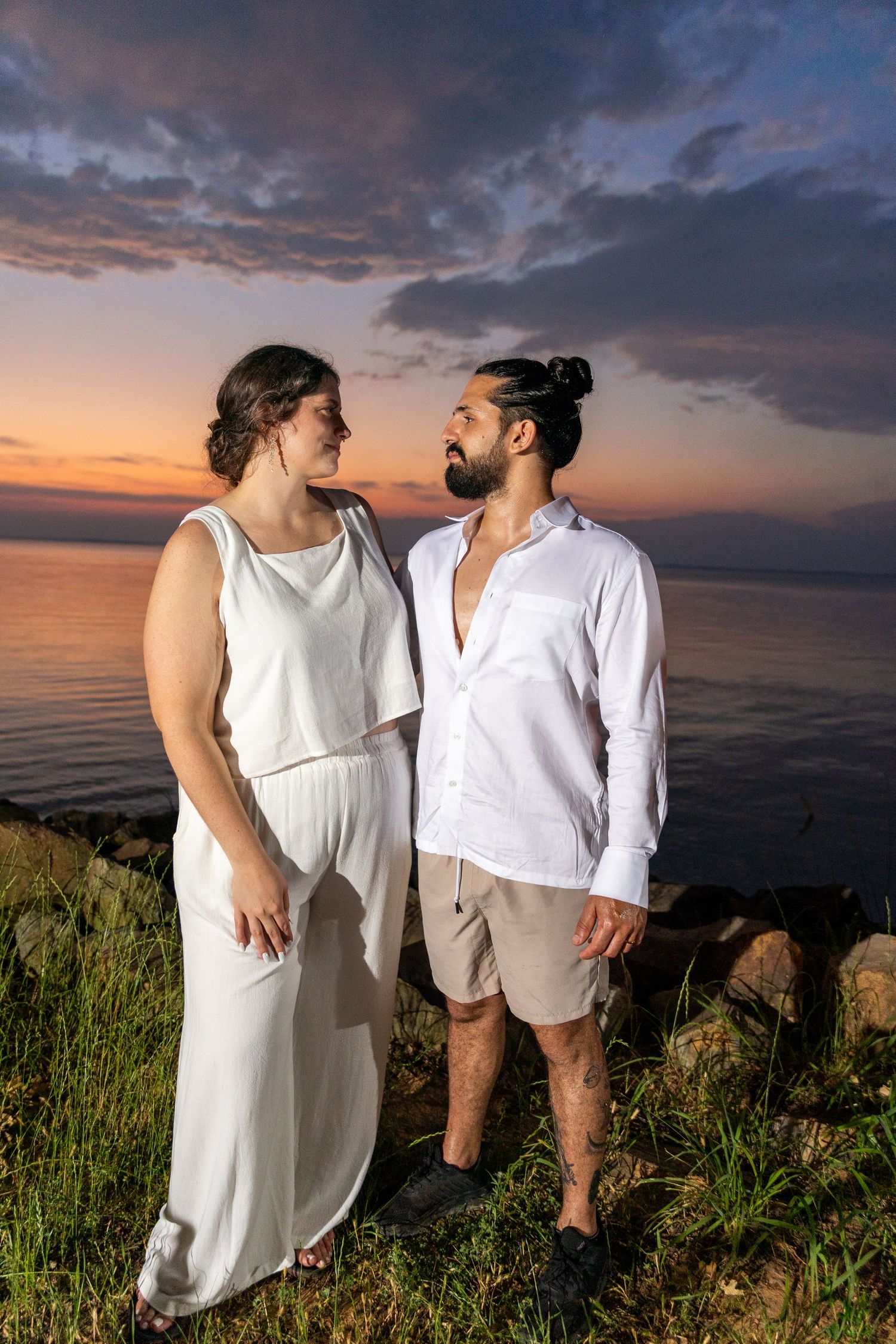 Couple in white outfits poses against dramatic sunset sky over ocean.