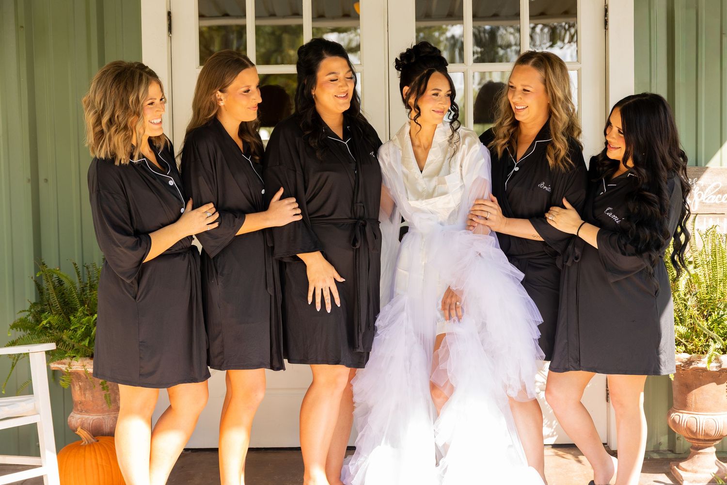 Bridesmaids in black dresses pose together on a porch during a wedding celebration.