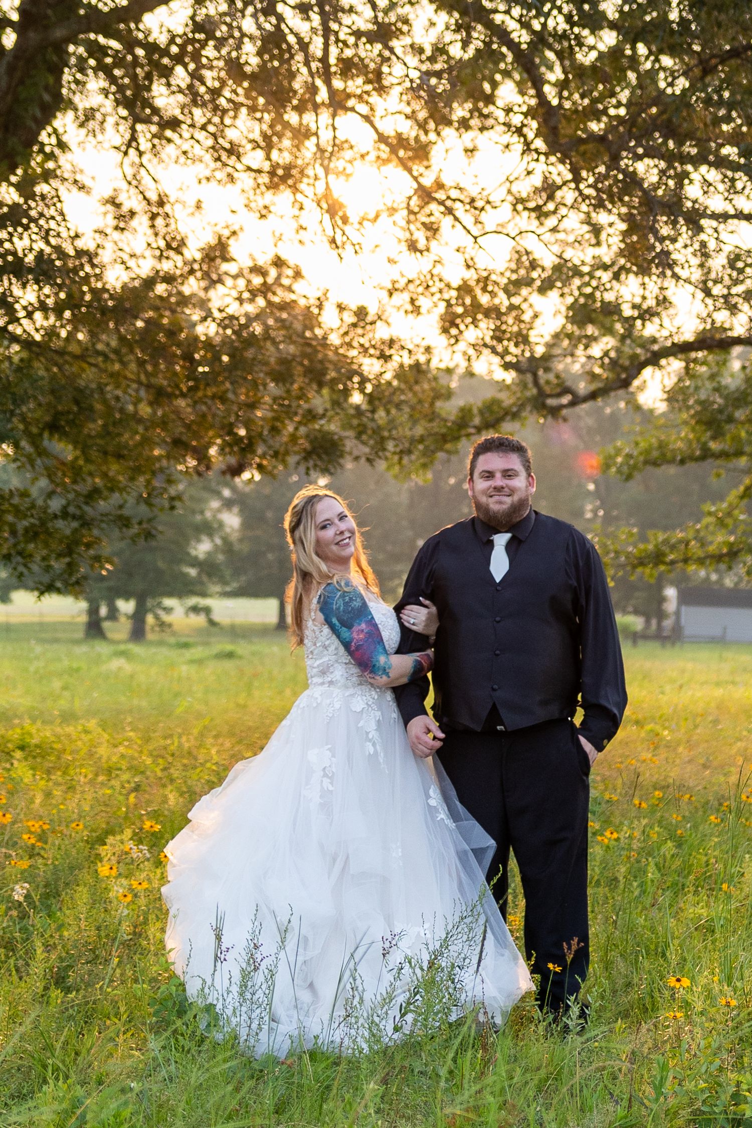 A bride and groom pose together in a sunlit meadow with golden wildflowers at sunset.
