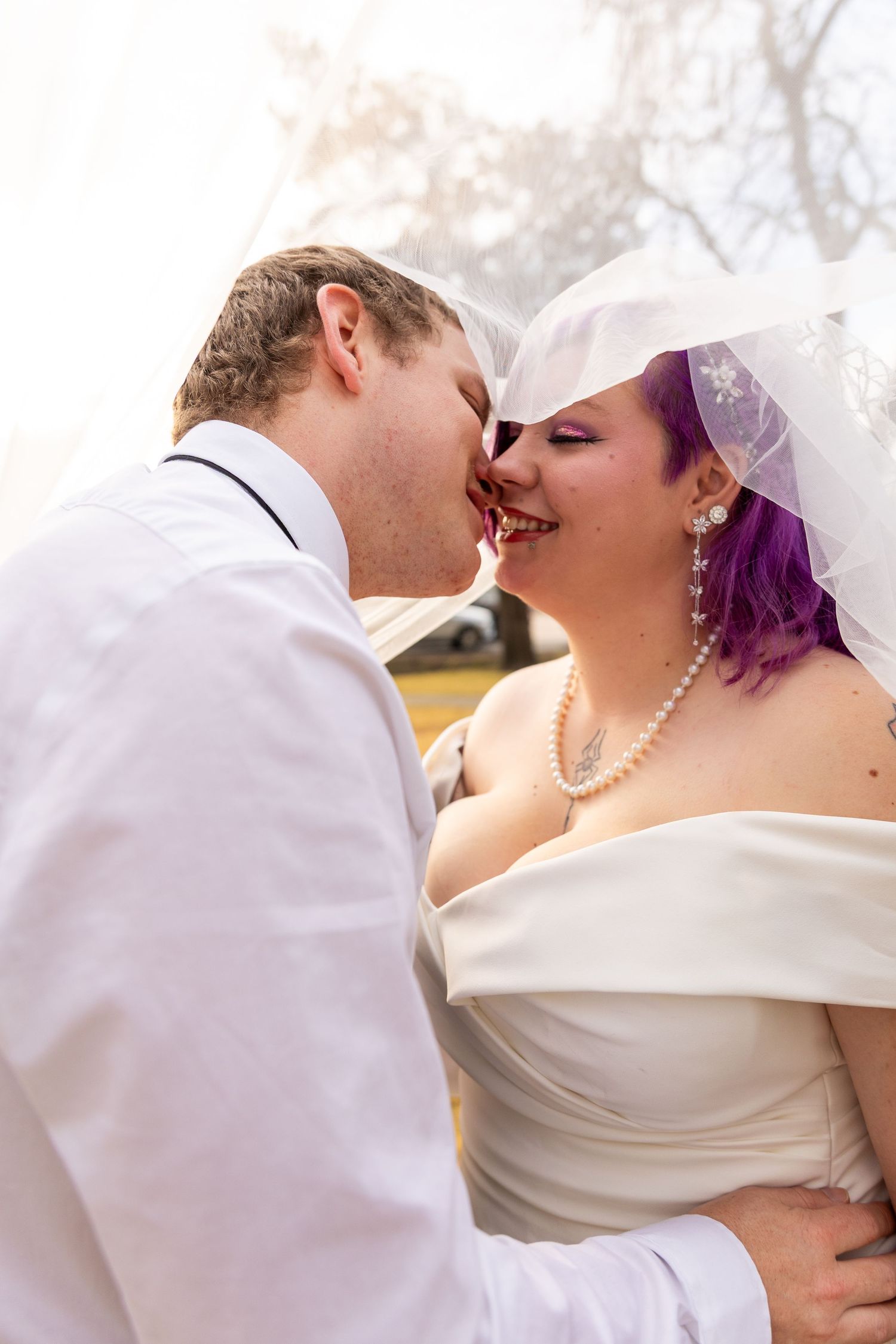 Bride with purple hair and groom sharing a kiss under a white veil on their wedding day.