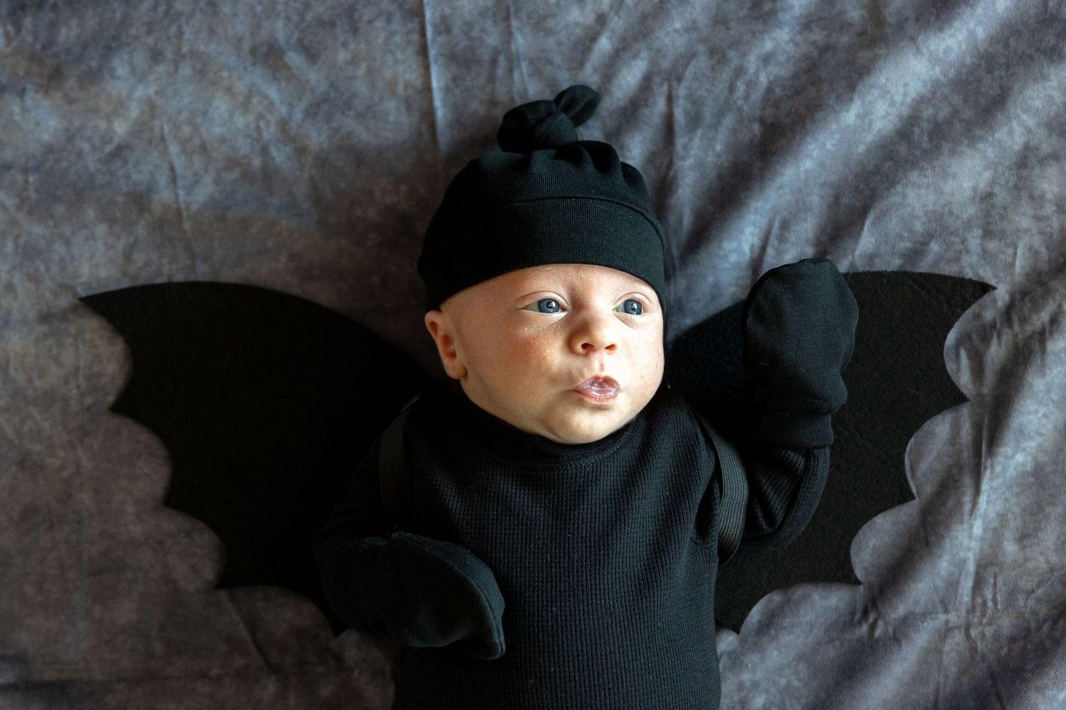 A baby wearing a black bat costume lying on dark fabric background.