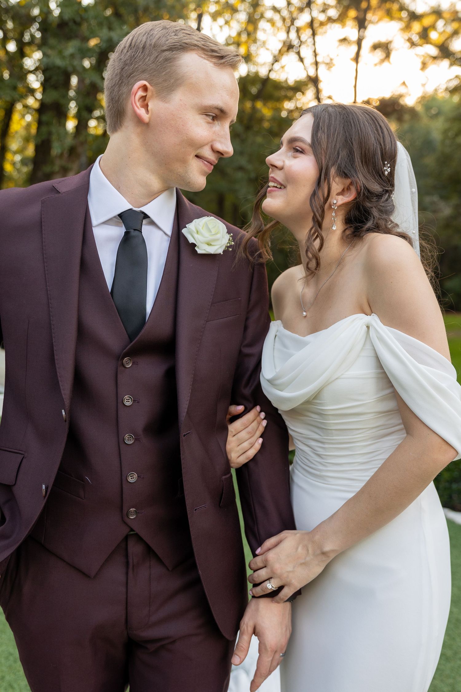 Romantic wedding portraits in a burgundy suit and white dress against natural outdoor backdrop.