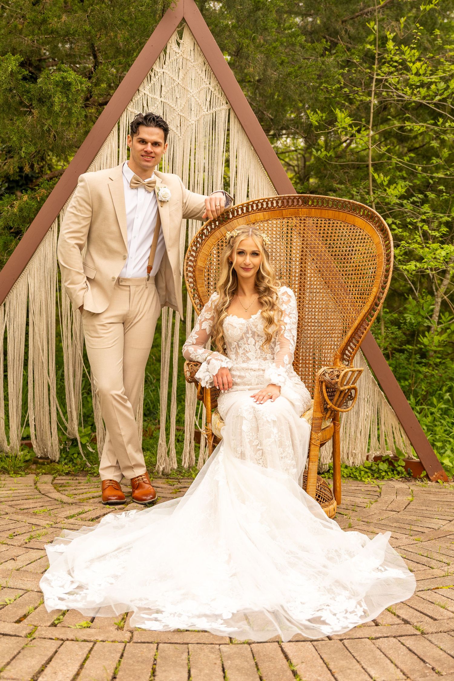 A wedding couple poses in front of a triangular macrame backdrop with a vintage peacock chair.