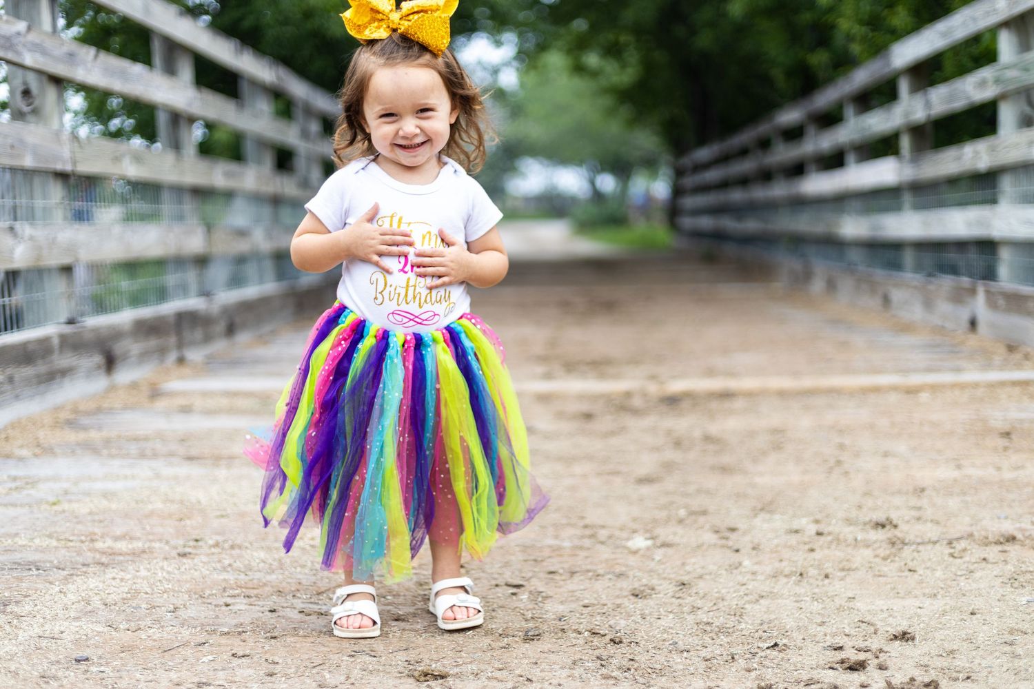 Young child in colorful tutu skirt and yellow bow playing on dirt path near white fence.