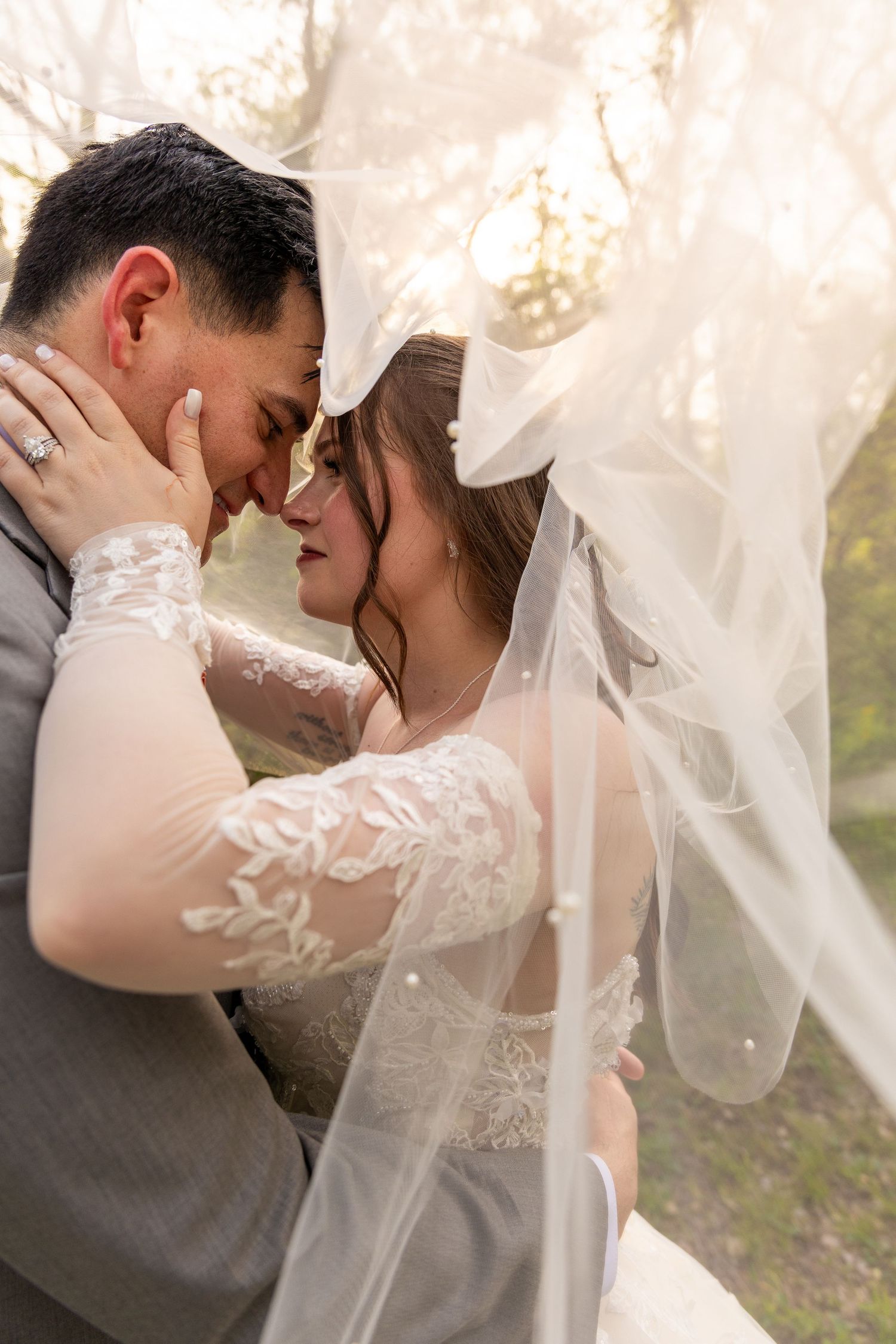 Romantic wedding couple sharing an intimate moment under flowing white veil in natural outdoor setting.
