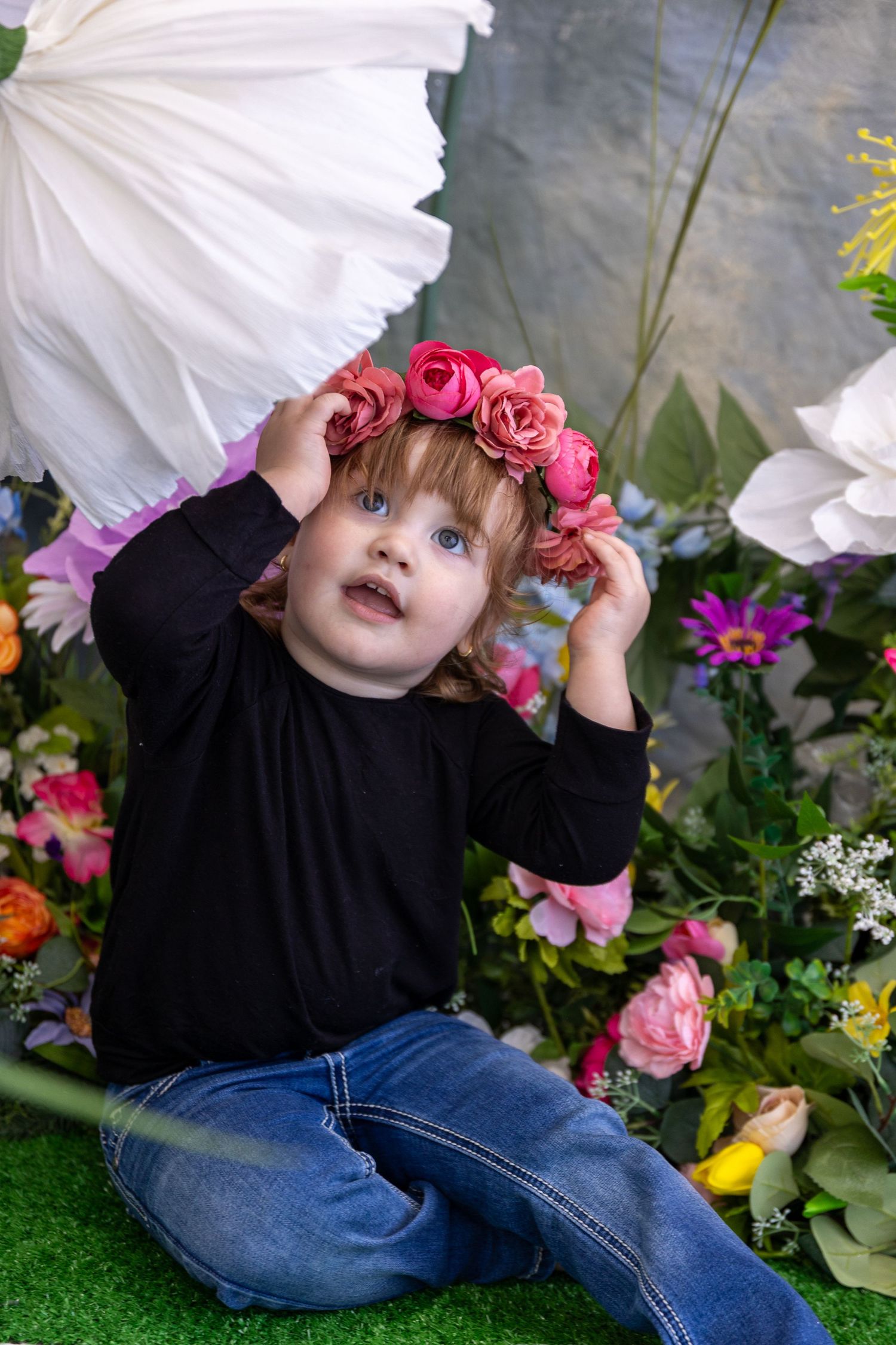 Child in black shirt poses playfully among white poppies and pink floral decorations.