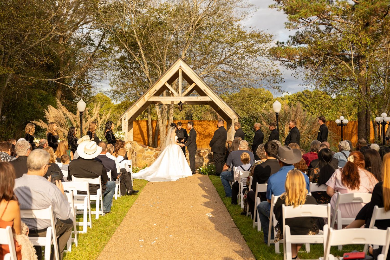 Outdoor wedding ceremony setup with wooden arch and rows of white chairs along tree-lined path.