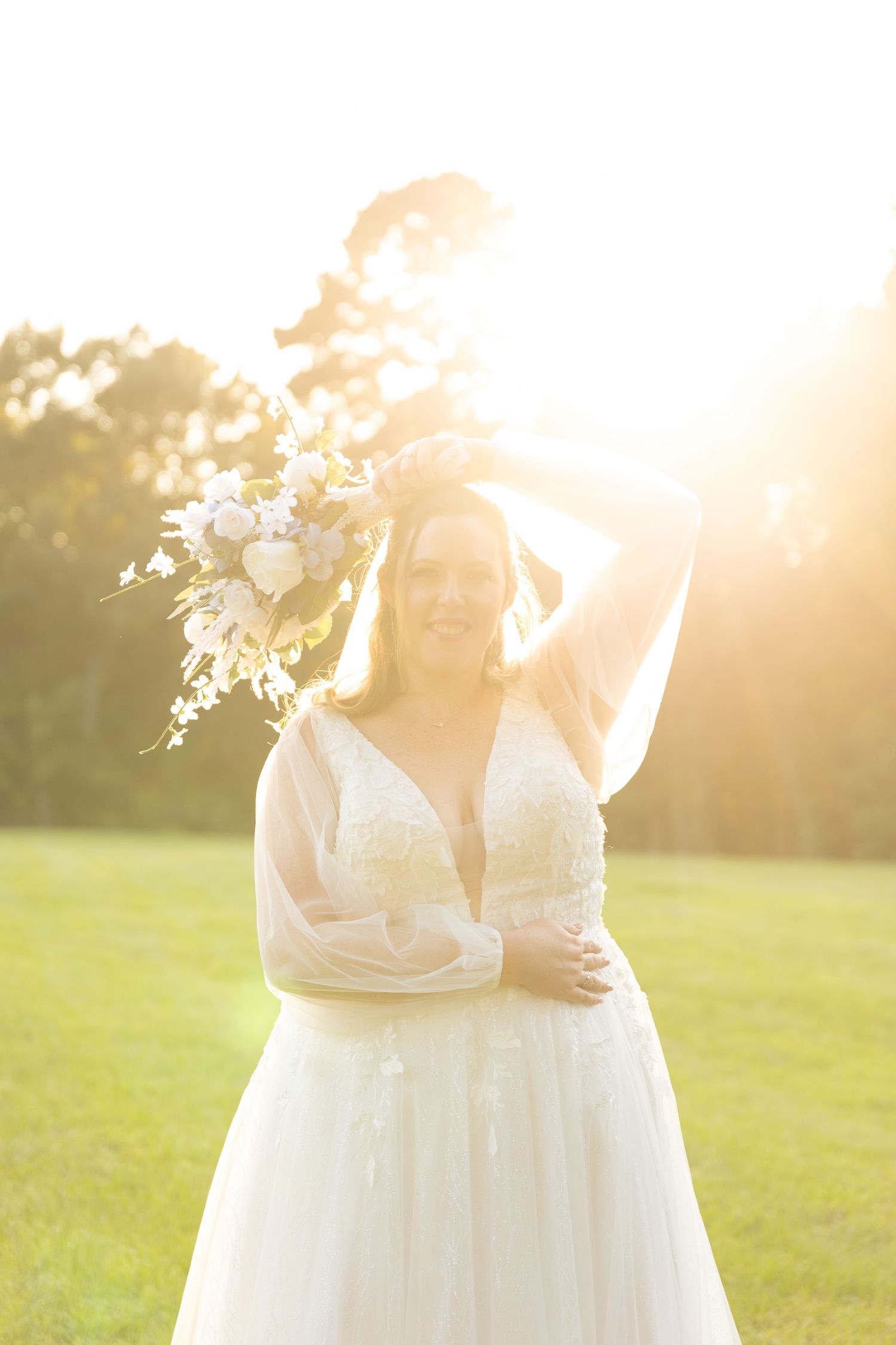 Bride in white lace gown holding floral bouquet overhead in golden sunset light on green lawn.