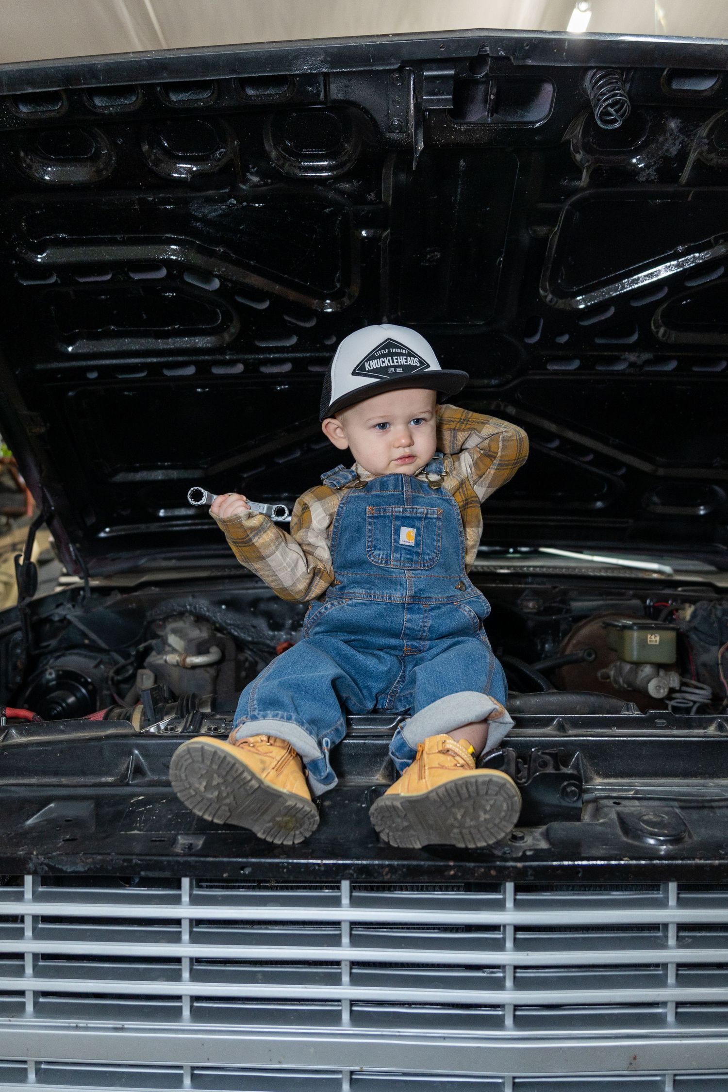 Small child in overalls and cap sits on engine compartment of vehicle during automotive photoshoot.