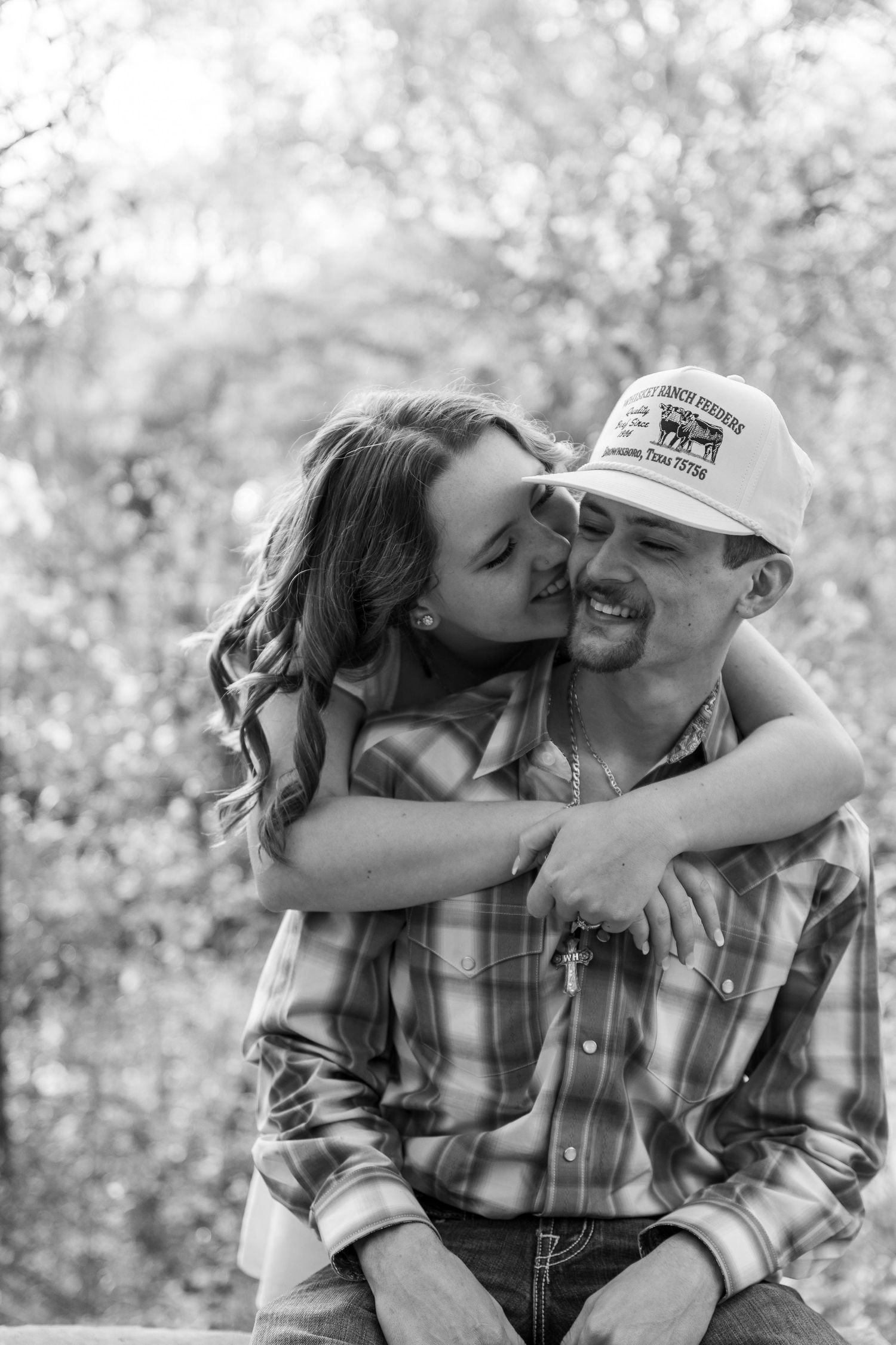 A black and white portrait of a couple sharing an intimate moment outdoors with one wearing a baseball cap.