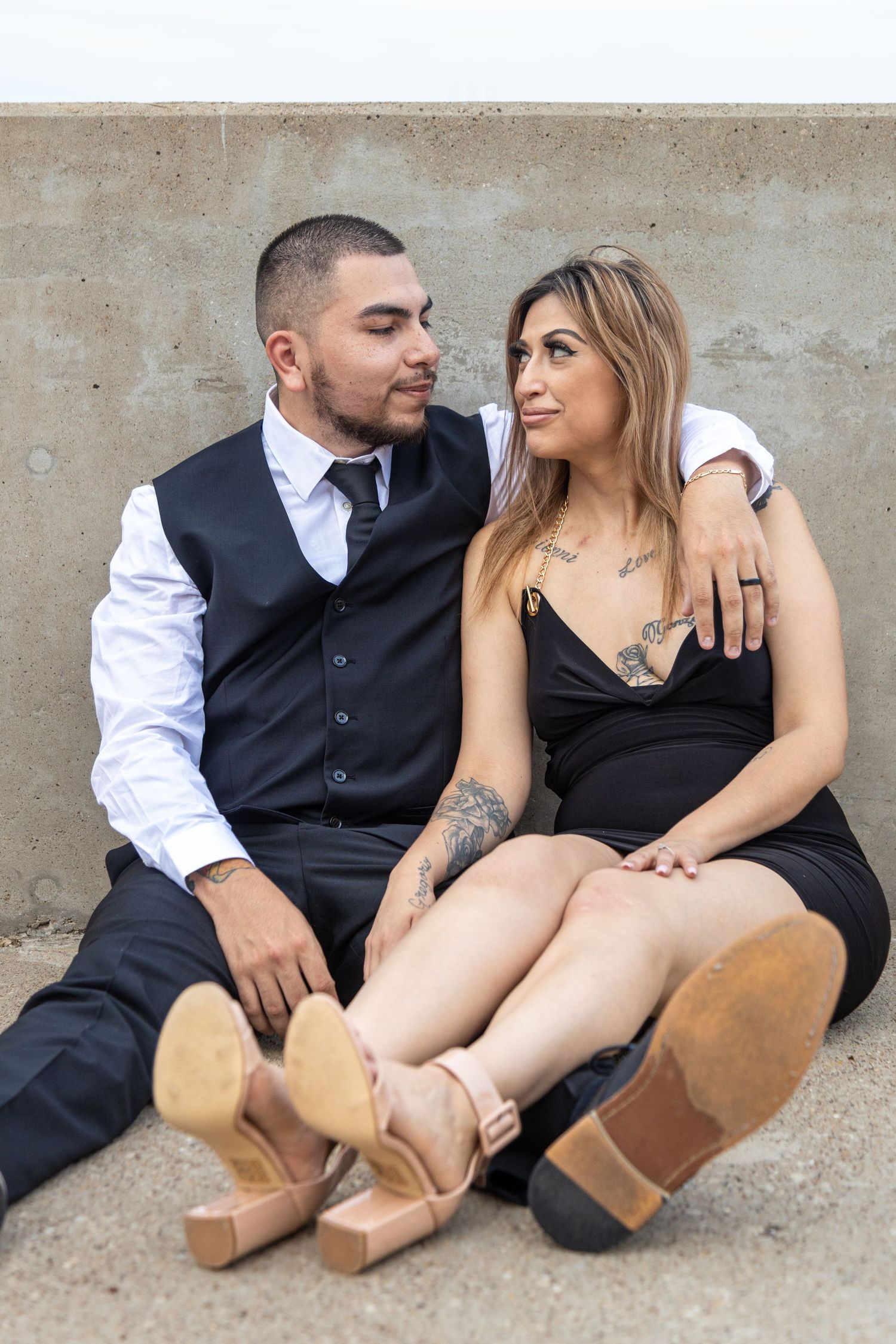 Couple in formal attire poses casually on concrete surface during urban photoshoot.