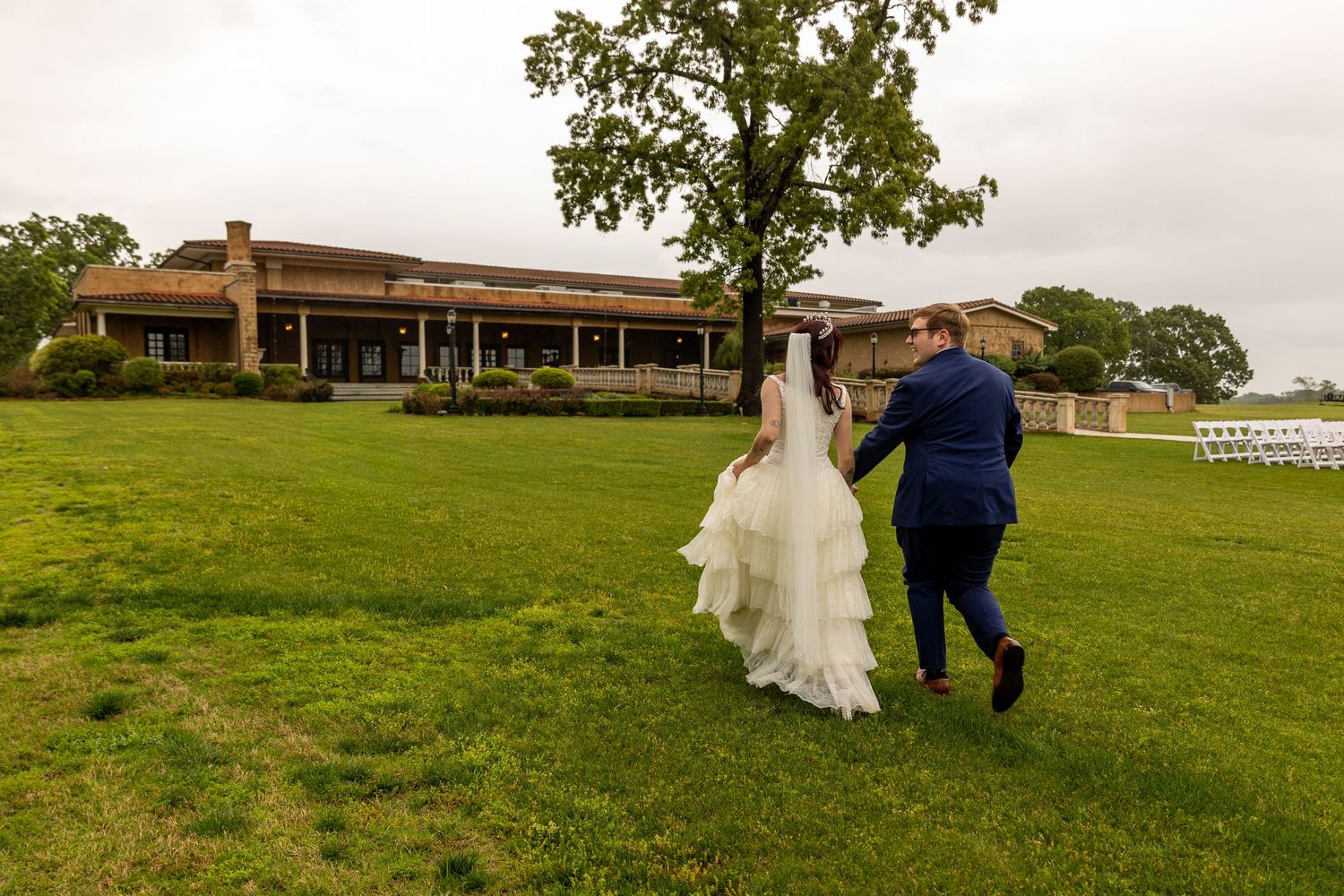 Bride and groom walking hand in hand across lush green lawn toward elegant wedding venue building.