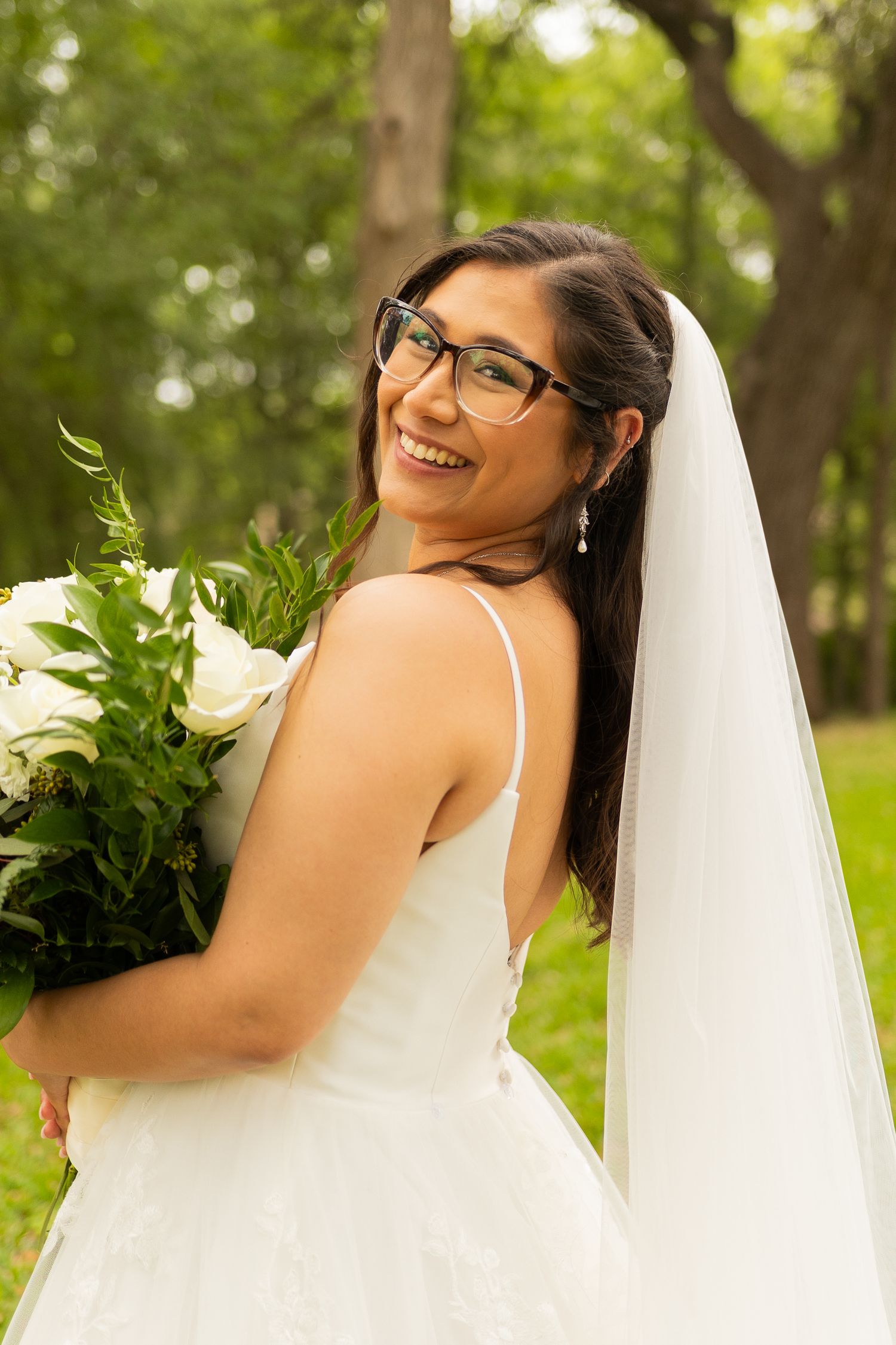 Bride in white dress and glasses poses with bouquet in outdoor garden setting.