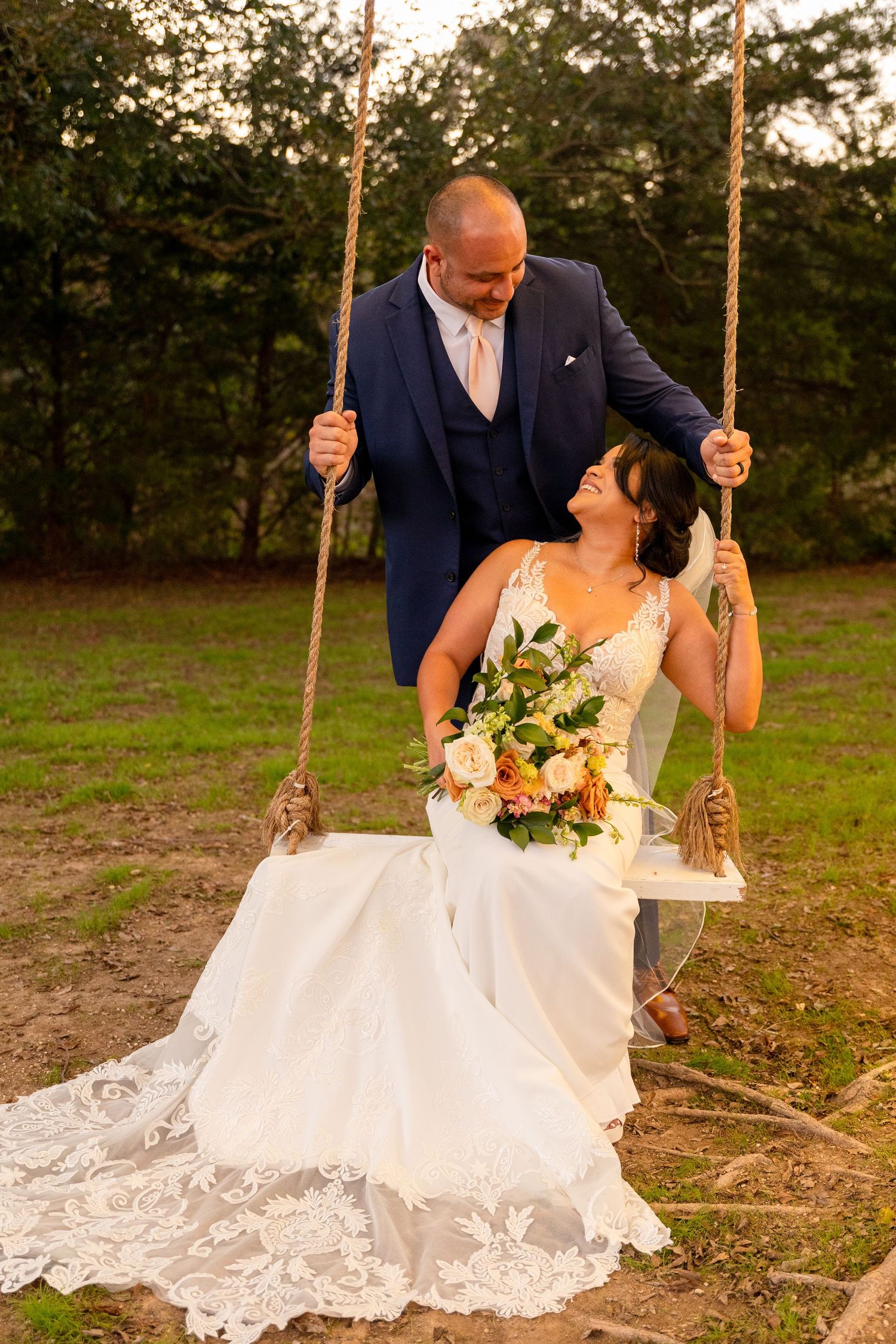 A bride in a lace dress sits on a wooden swing during sunset wedding photos.