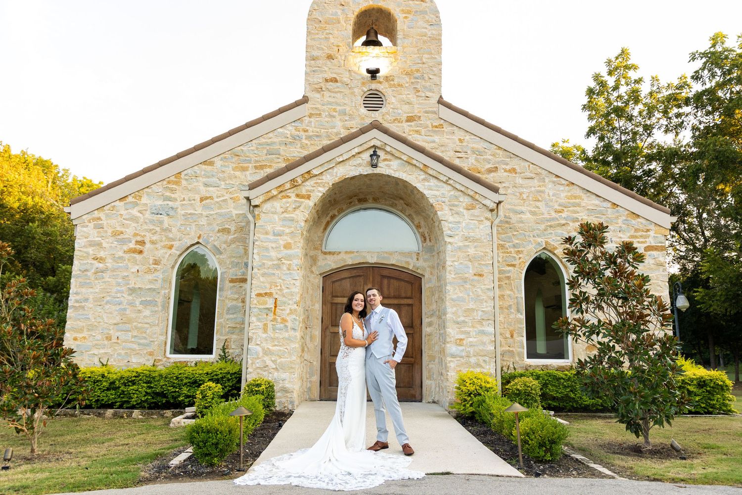 Wedding couple poses in front of rustic stone chapel with elegant architectural details.