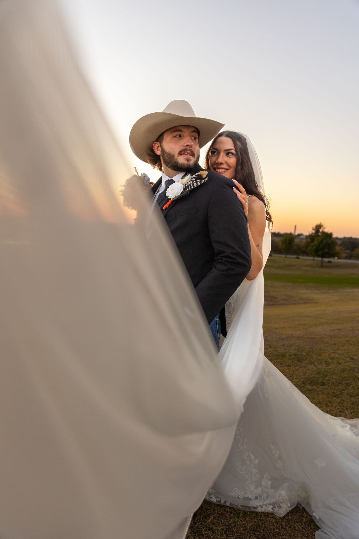 A couple shares romantic moments at sunset in a field with flowing white wedding dress and cowboy hat.