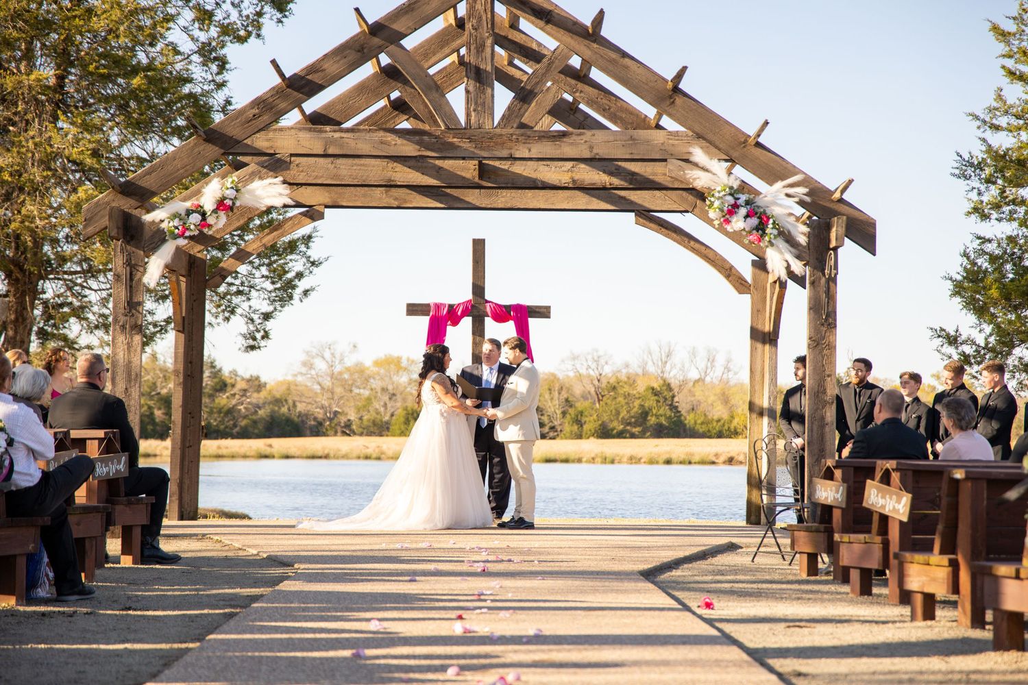 Outdoor wedding ceremony under a wooden pergola with guests seated on either side.
