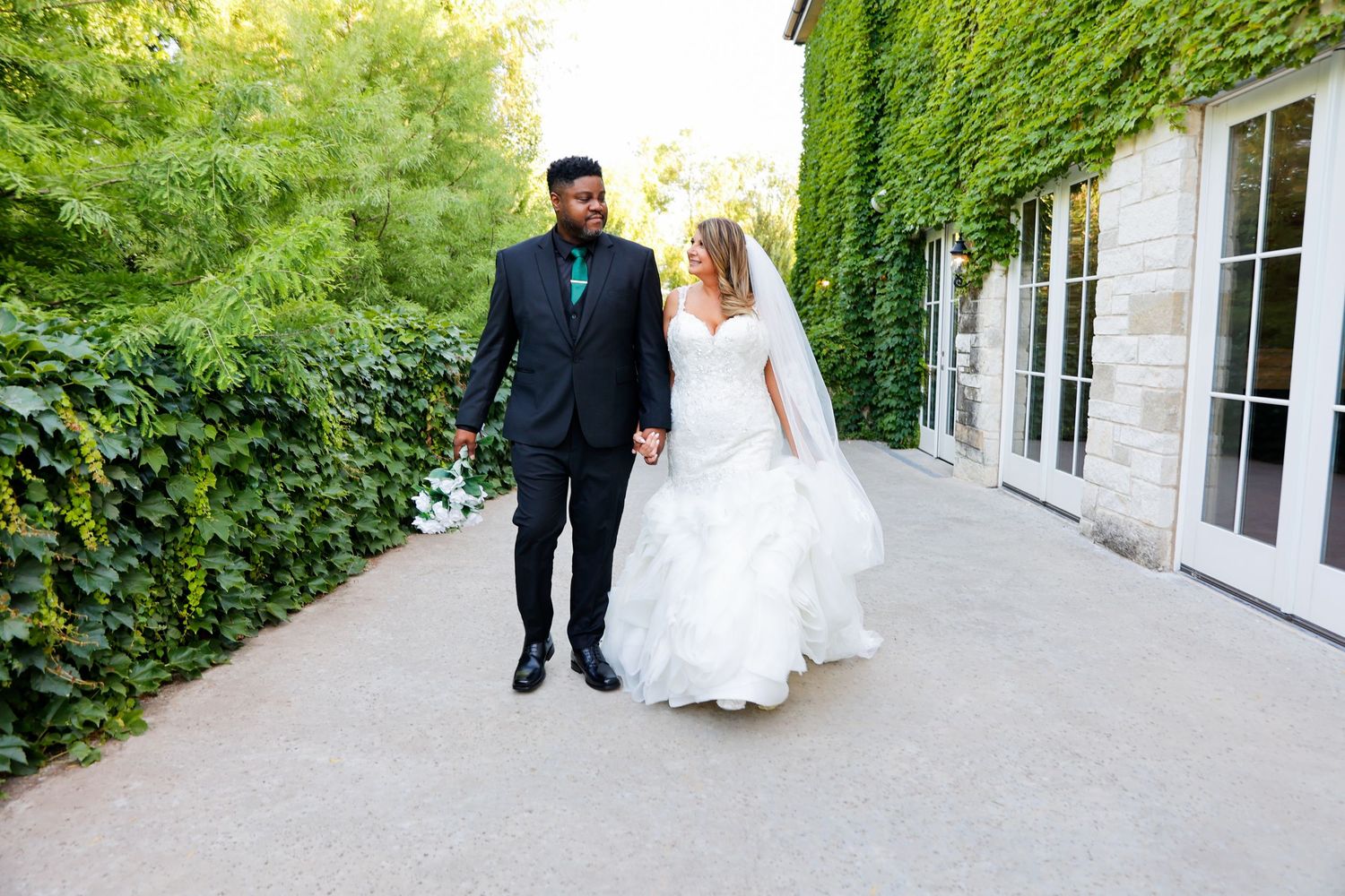Wedding couple walking along ivy-covered building exterior in natural light.