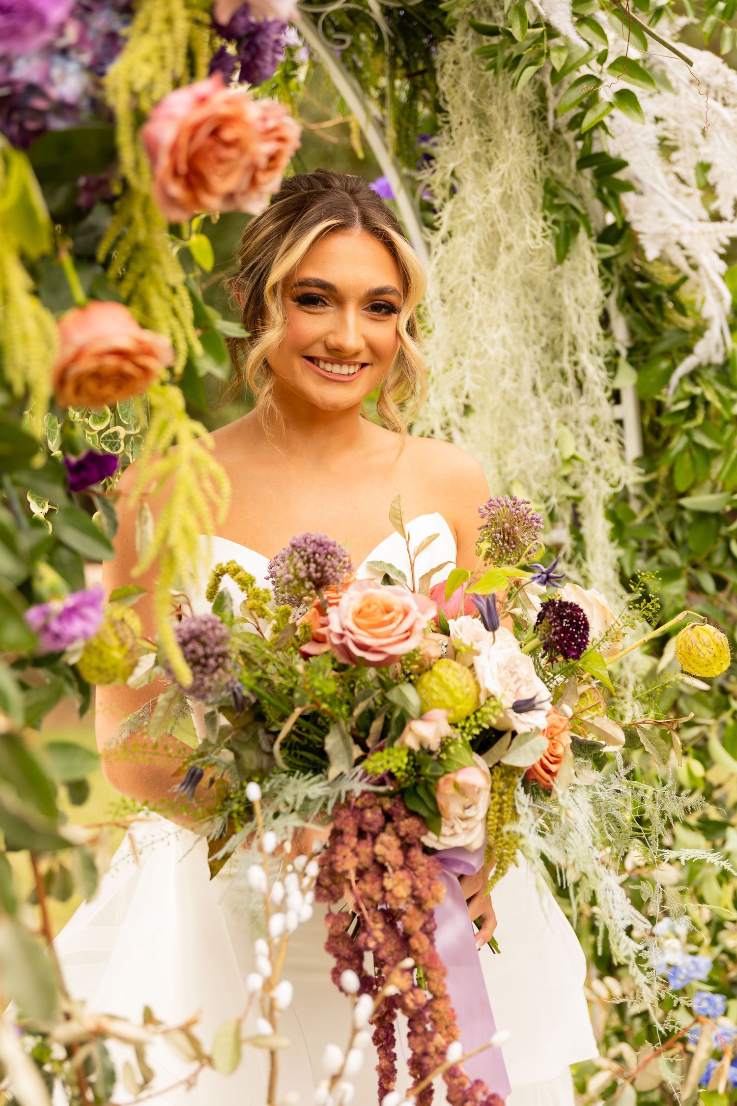 Bride with floral bouquet poses under decorative arch with hanging flowers and greenery.